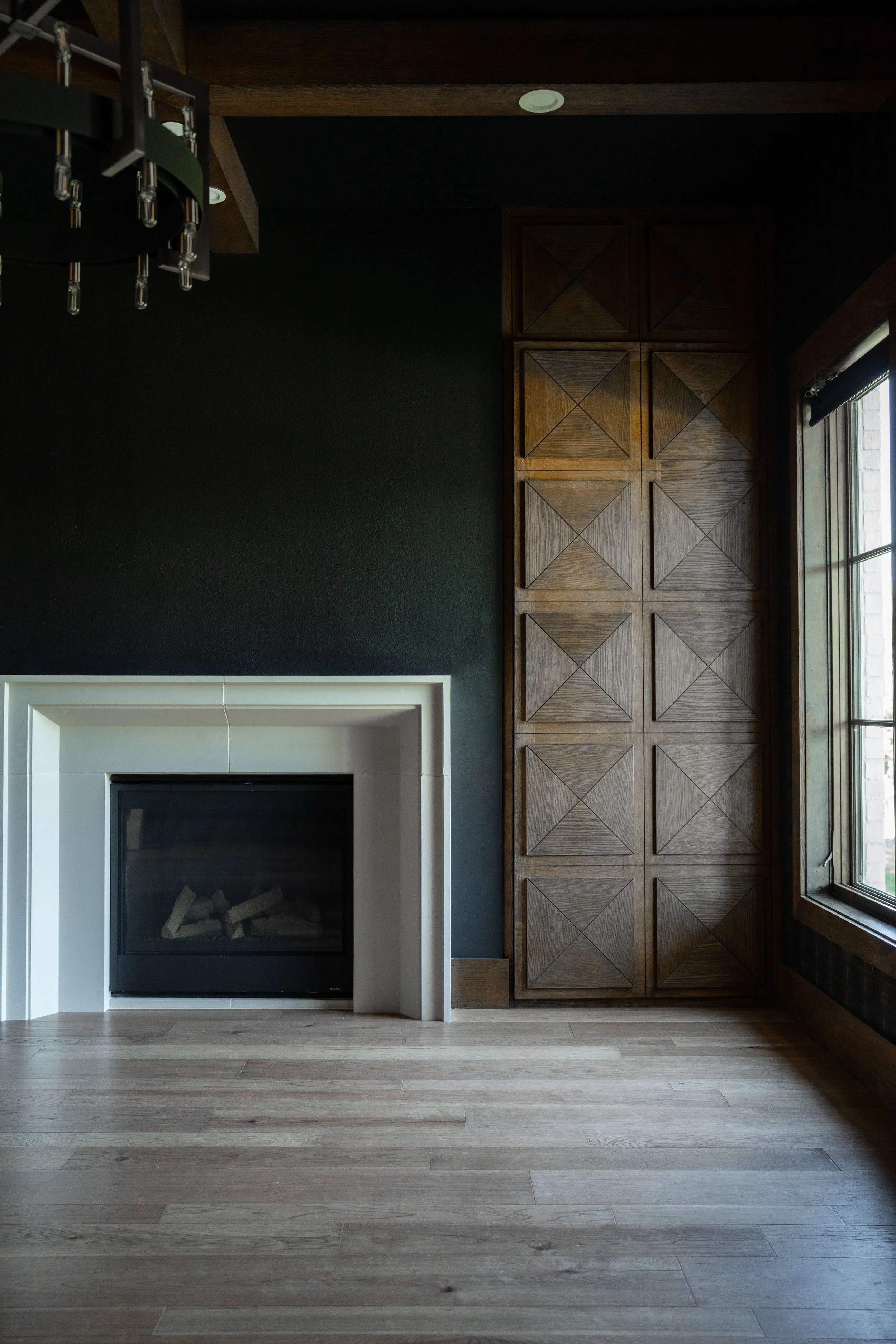 Interior of a room featuring a fireplace with a white mantel, a dark green wall, wooden flooring, and a large window with wooden blinds, with built-in wooden cabinets on the right side.