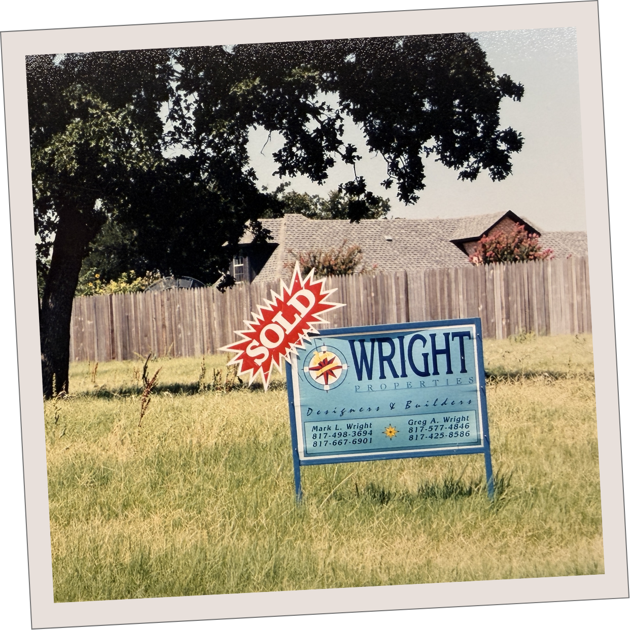 An old real estate for sale sign with 'SOLD' label in front of a grassy yard, wooden fence, and house with trees in the background.
