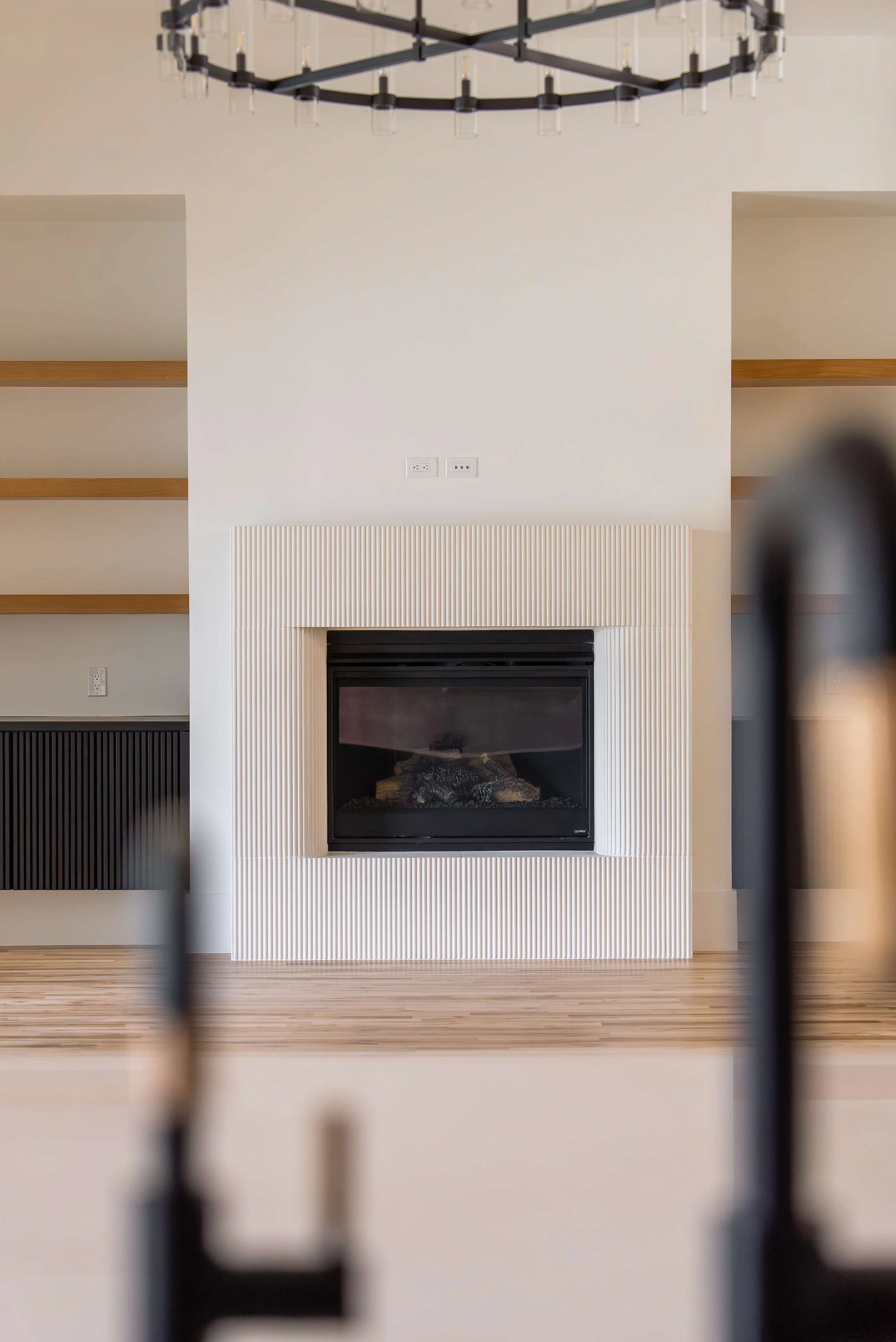 Luxury living room in Keller, TX with a white textured fireplace, light wood floor, and floating wooden shelves on either side of the fireplace. A modern black-and-white chandelier hangs from the ceiling.