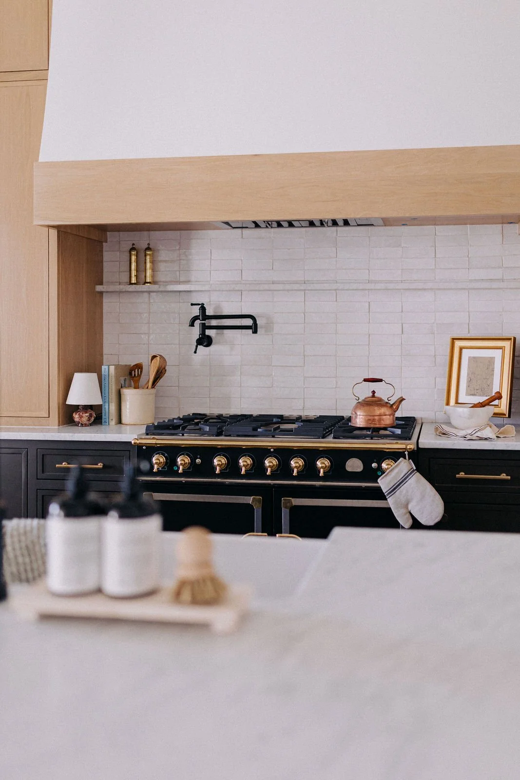 Modern kitchen with black and gold stove, copper kettle, framed artwork, white brick backsplash, and various kitchen items on the counter.