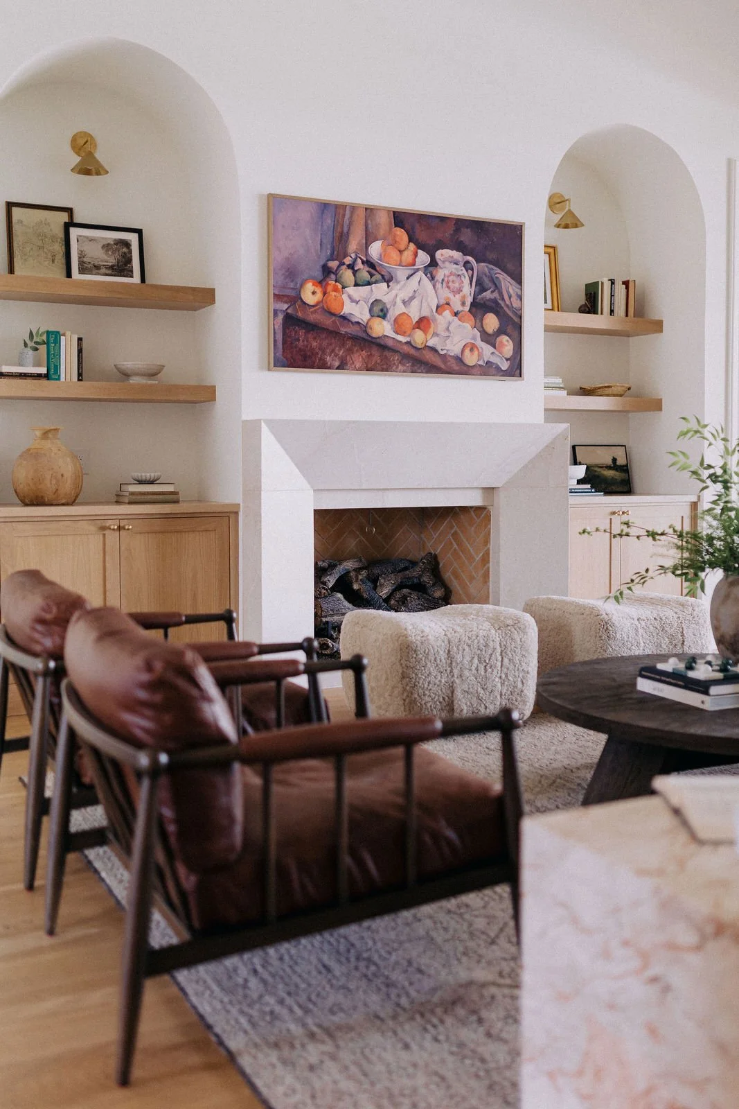 A cozy living room with a fireplace, a painting of fruit above the mantel, built-in shelves with books and decorative items, leather armchairs with wooden frames, and plush ottomans.