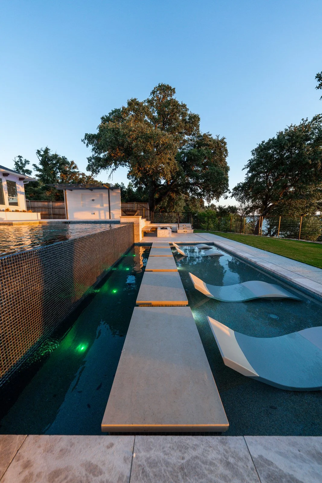 Modern backyard swimming pool with floating lounge chairs, trees, and a clear sky during sunset.
