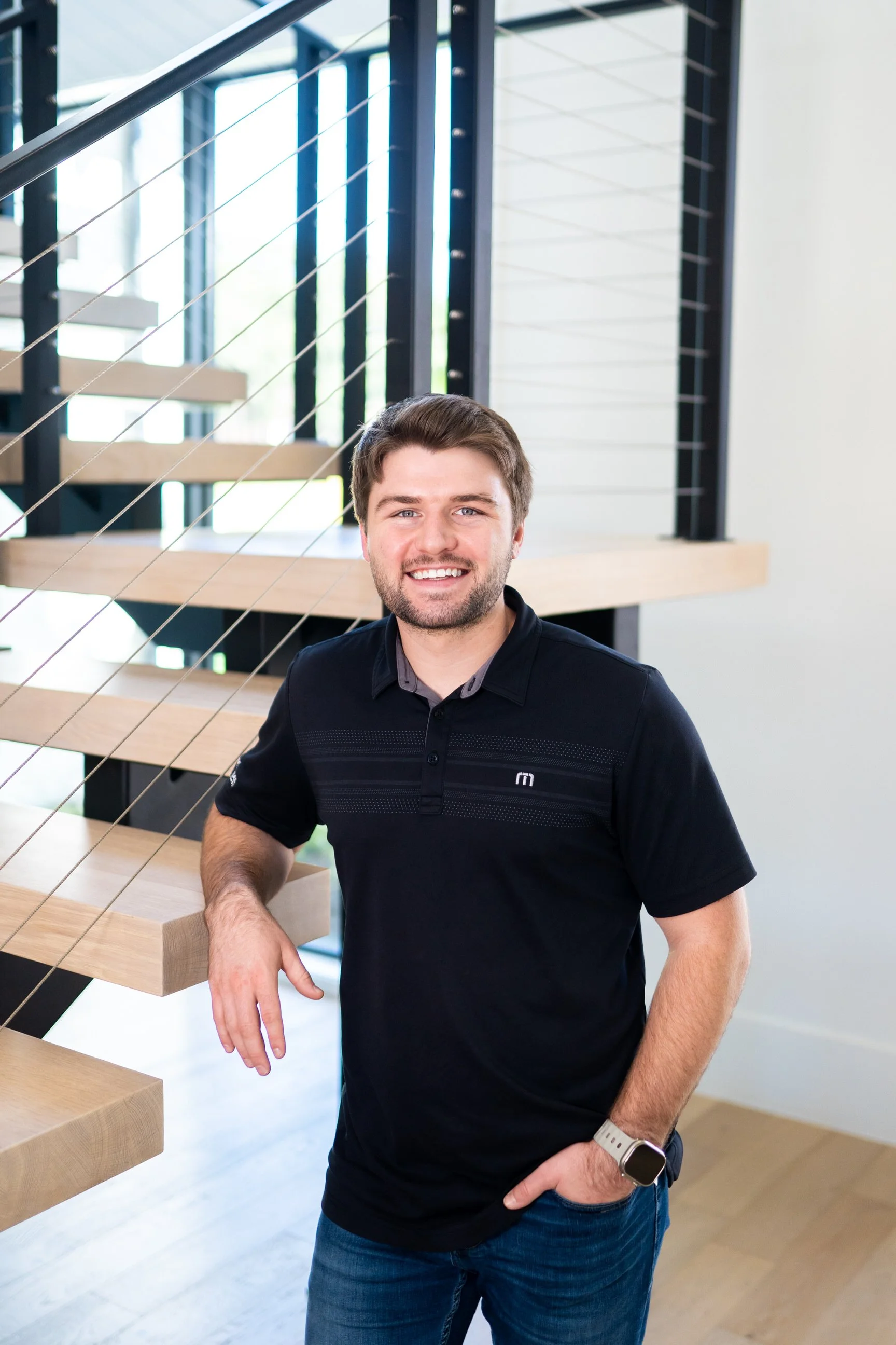 A young man with brown hair, beard, and a broad smile, wearing a black polo shirt and blue jeans, standing beside a modern staircase with wooden steps and metal railing in a bright, contemporary interior.