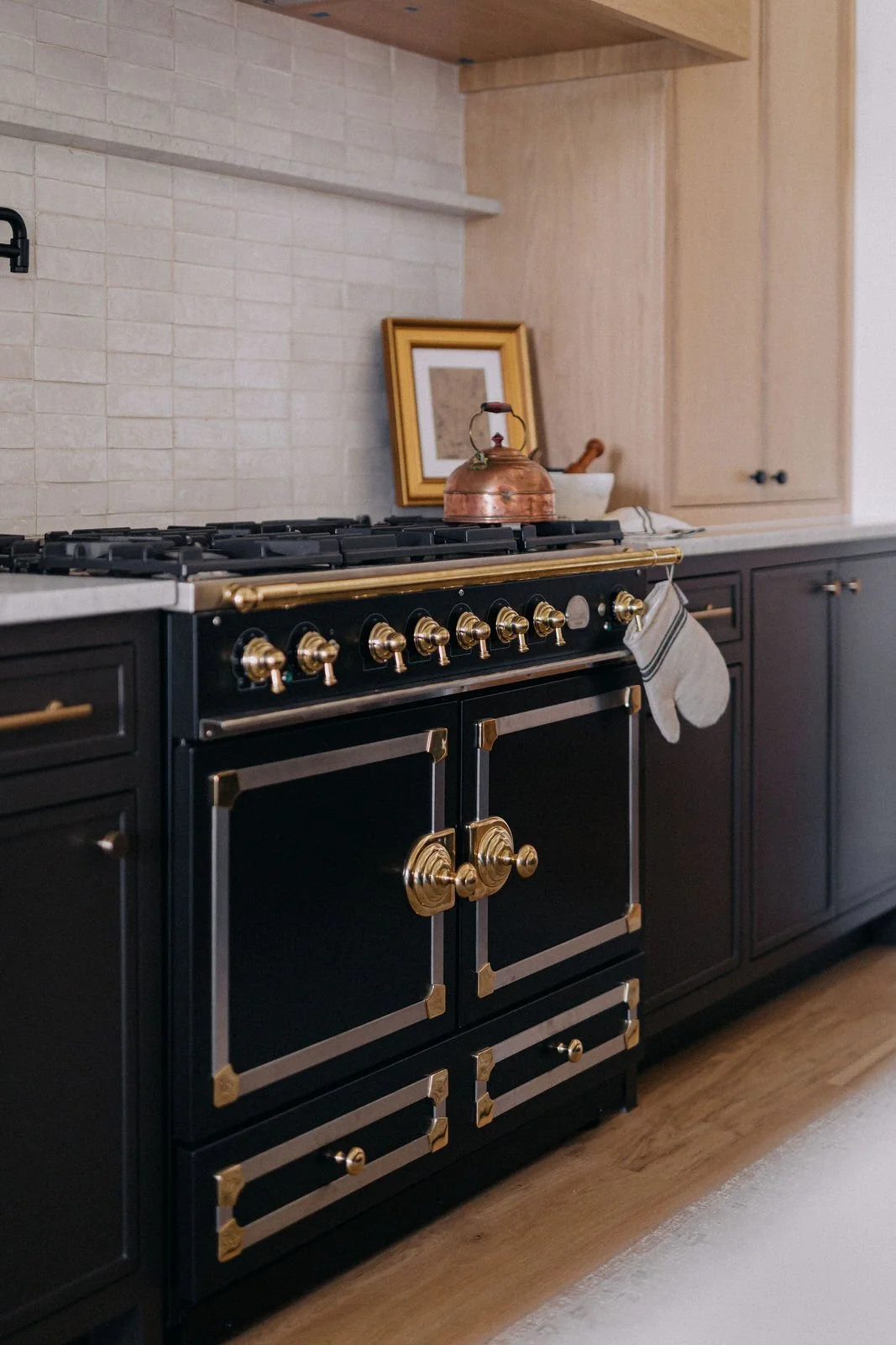 A black and gold vintage style stove in a modern kitchen with dark cabinetry, wooden flooring, a beige tile backsplash, a copper kettle, a picture frame, and a white dish.
