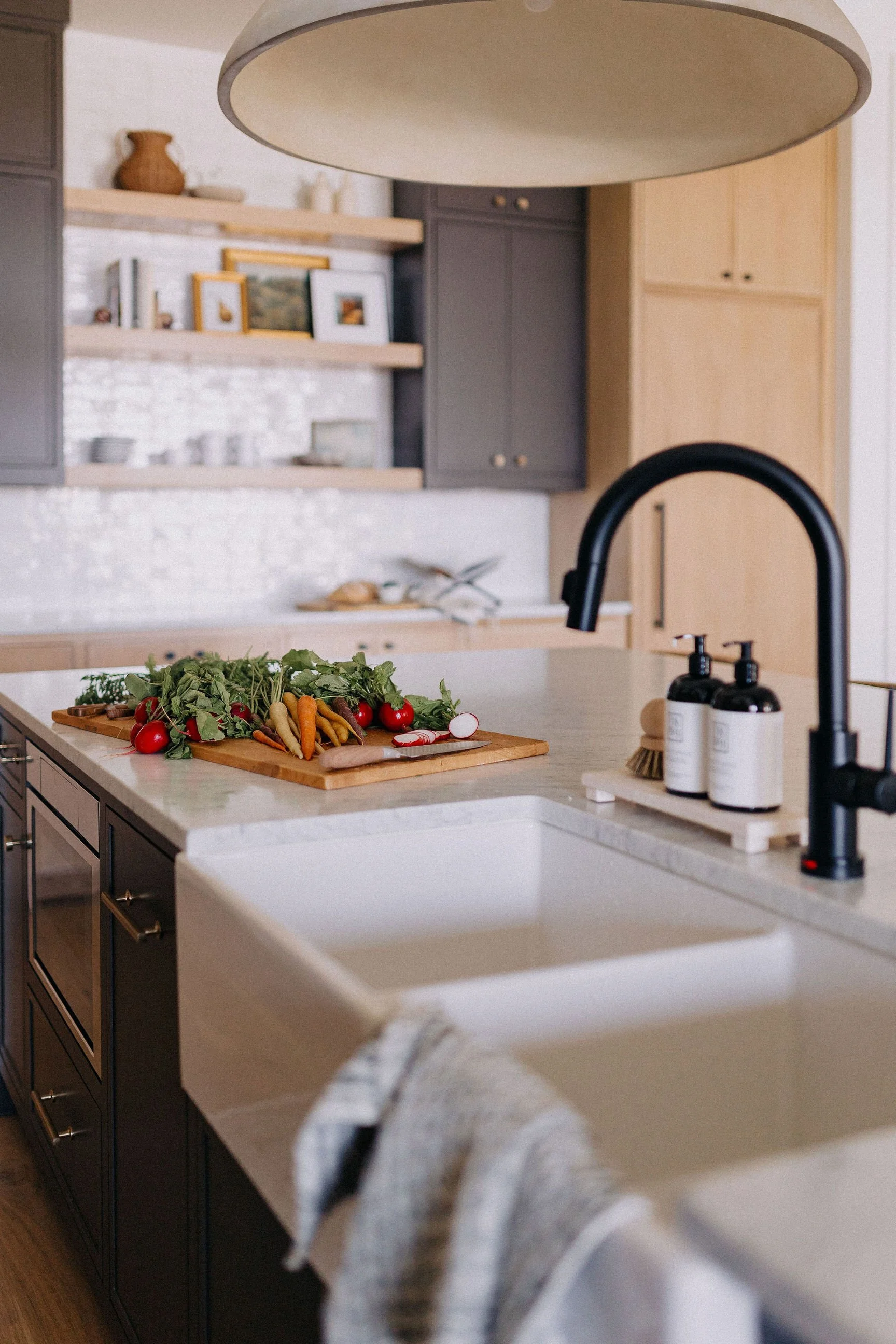 A modern kitchen with a white island countertop, dark cabinet drawers, and a black sink faucet. Fresh vegetables, including carrots, radishes, and tomatoes, are on a wooden cutting board on the counter. Open shelving with decorative items and a large ceiling light are also visible.