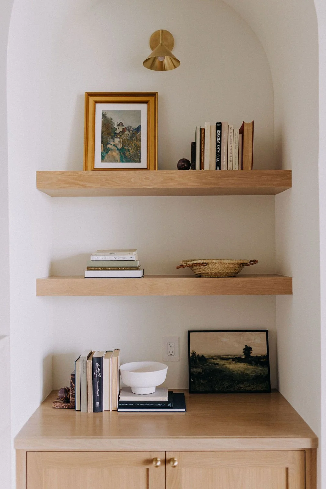 Wooden shelves with books, a framed landscape picture, a white bowl, and decorative objects on a wooden cabinet against a white wall.