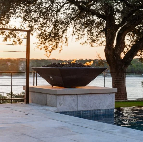 Luxury, modern outdoor fire bowl on a stone platform overlooking a river at sunset, with trees and a metal railing in the background.