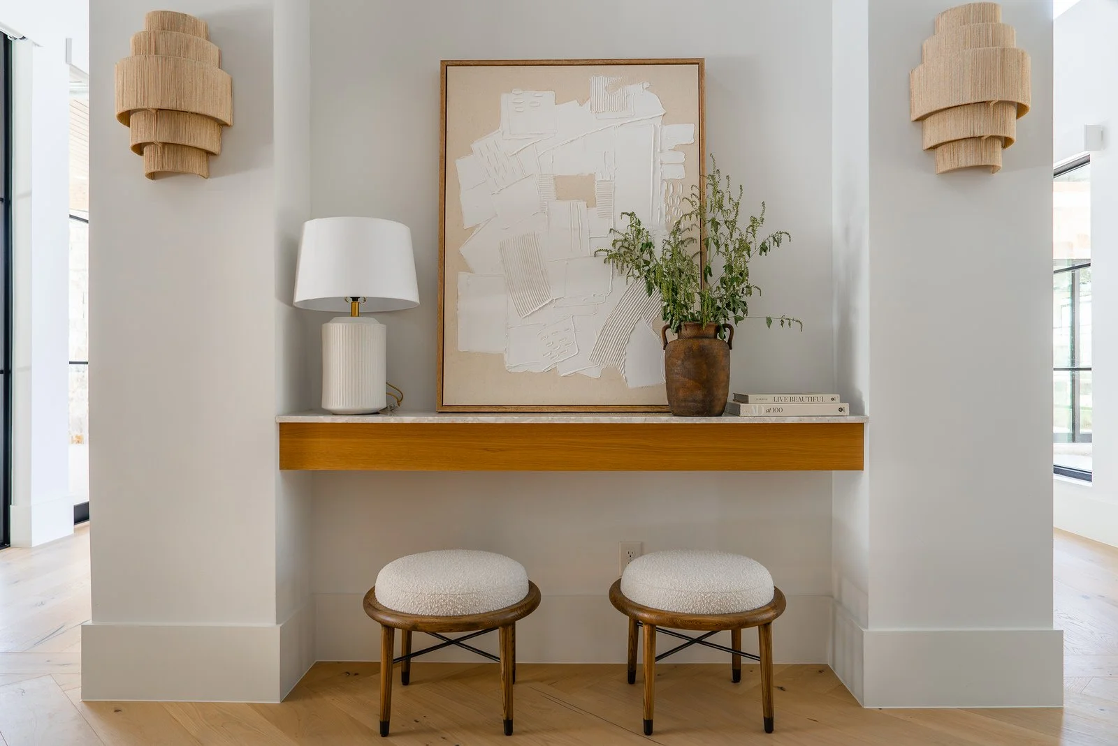 Interior space with a floating wooden shelf, two beige upholstered stools, a white table lamp, a framed abstract artwork, a potted plant, and books, in a modern minimalist style.