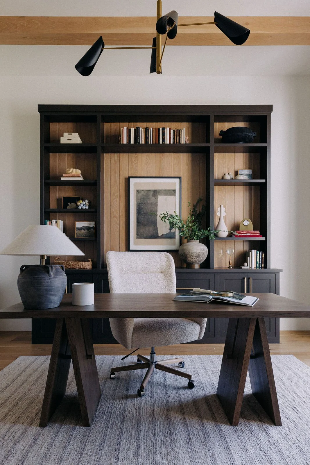 Modern home office with a wooden desk, beige upholstered swivel chair, black and wood bookshelf with decorative items and books, abstract artwork, and a black and beige table lamp on a textured rug.