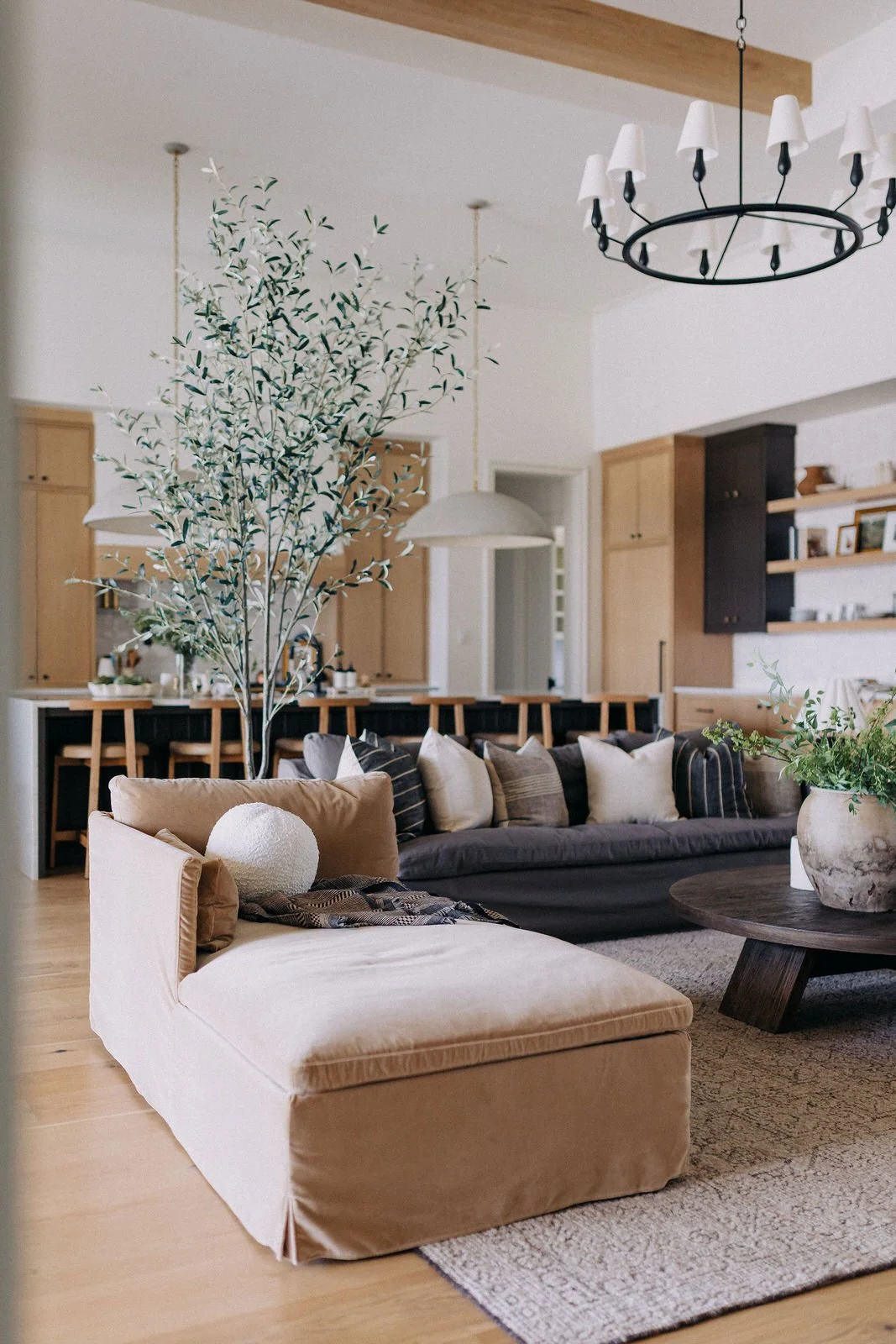 A living room with beige and dark brown couches, a round wooden coffee table, and large potted plants. The background features a kitchen with light wood cabinets and a bar with black and wooden chairs. A chandelier hangs from the ceiling.
