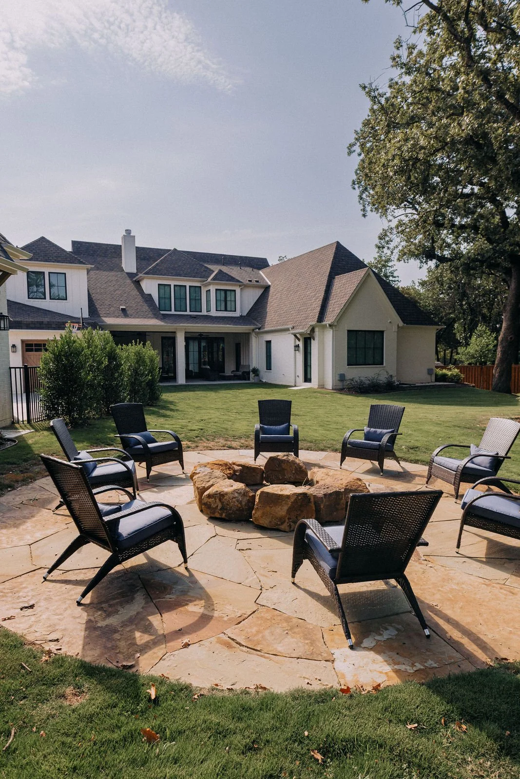 Backyard with a circular arrangement of six black chairs around a fire pit made of large rocks, on a stone patio, with a well-maintained lawn, trees, and a white house with a gabled roof in the background.