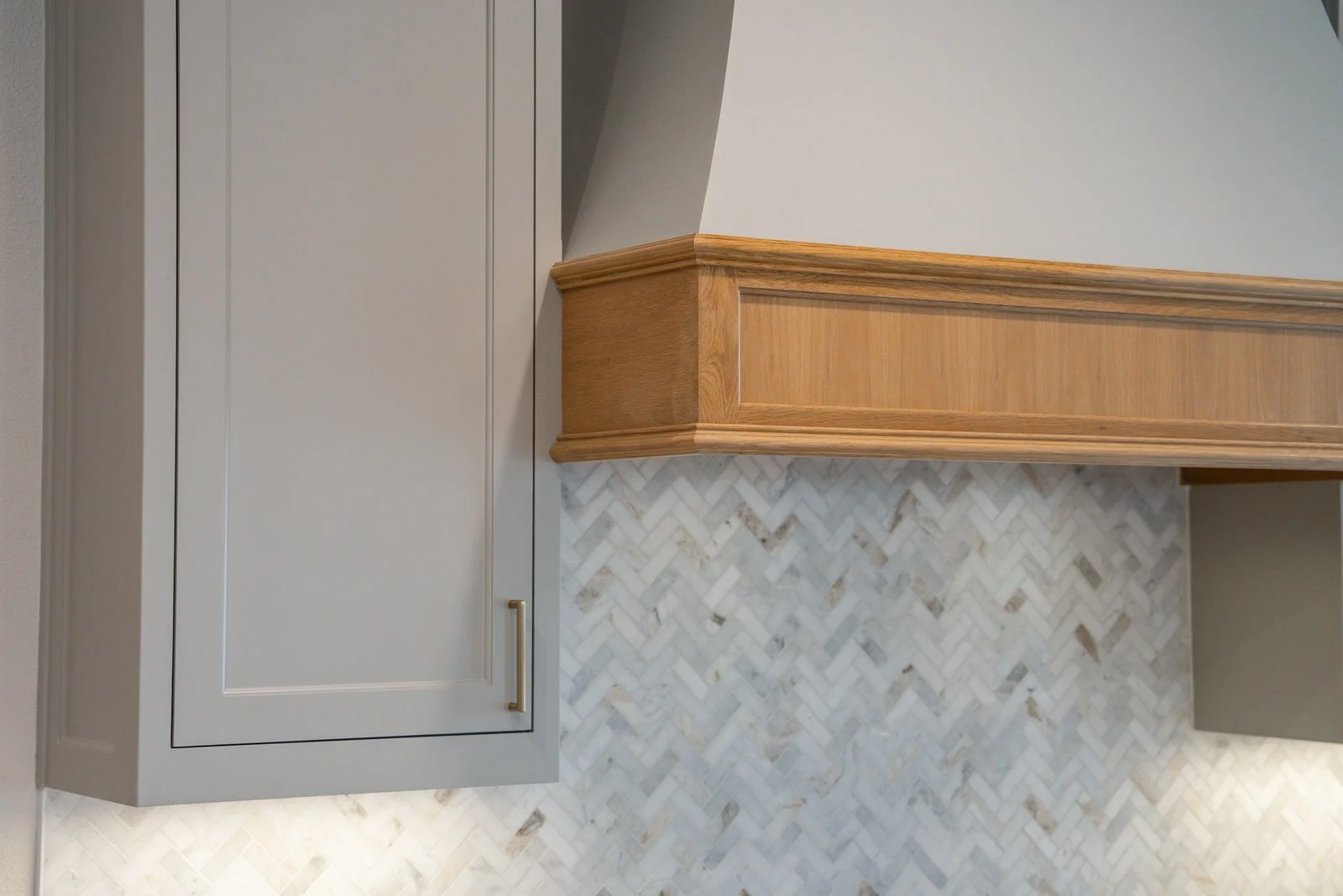 Close-up view of custom kitchen cabinetry with a mix of light grey cabinet and wooden trim, along with a herringbone patterned marble backsplash.