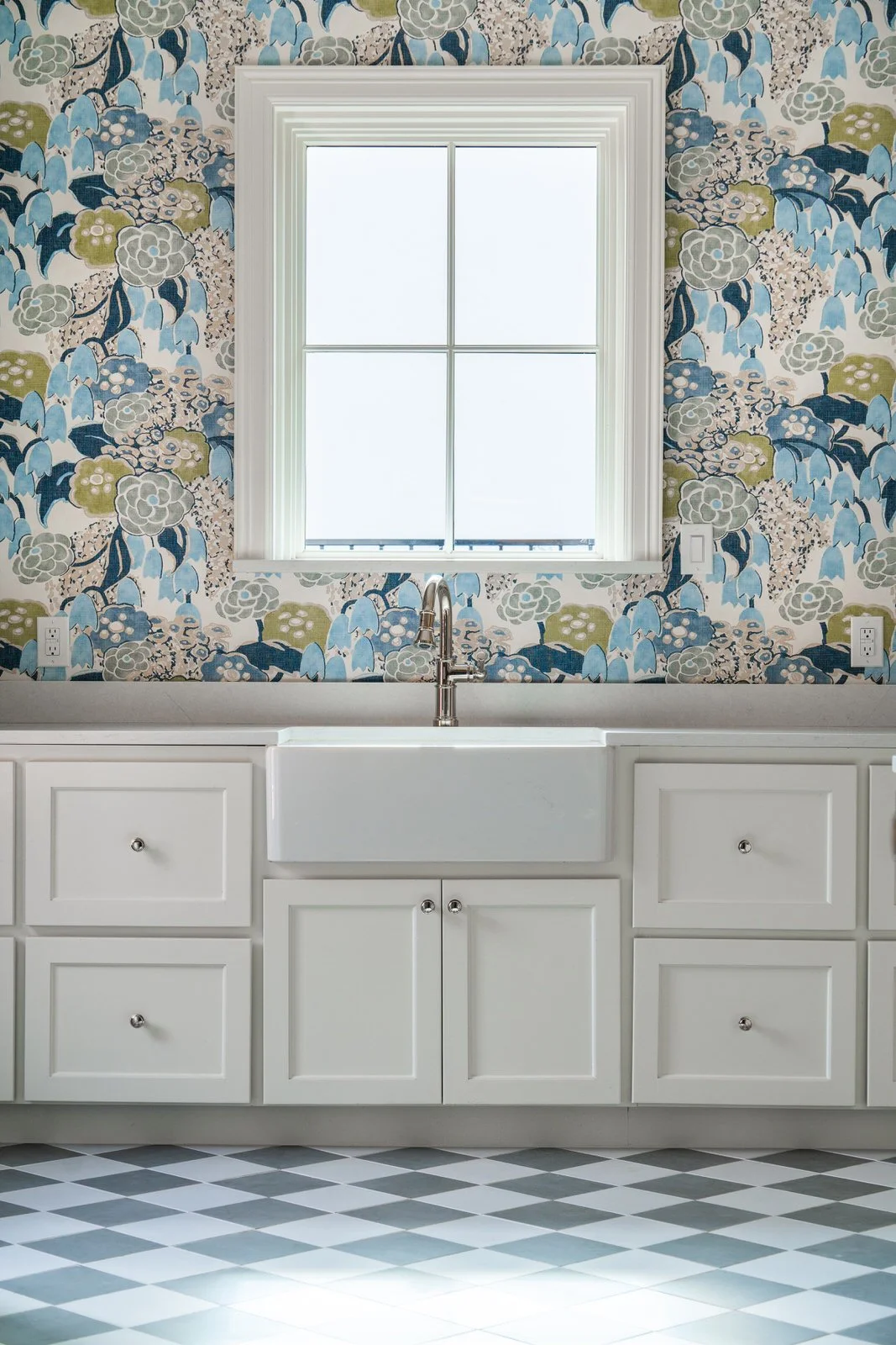 Kitchen with white cabinets, a farmhouse sink, and a window with floral wallpaper in shades of blue, gray, and green.