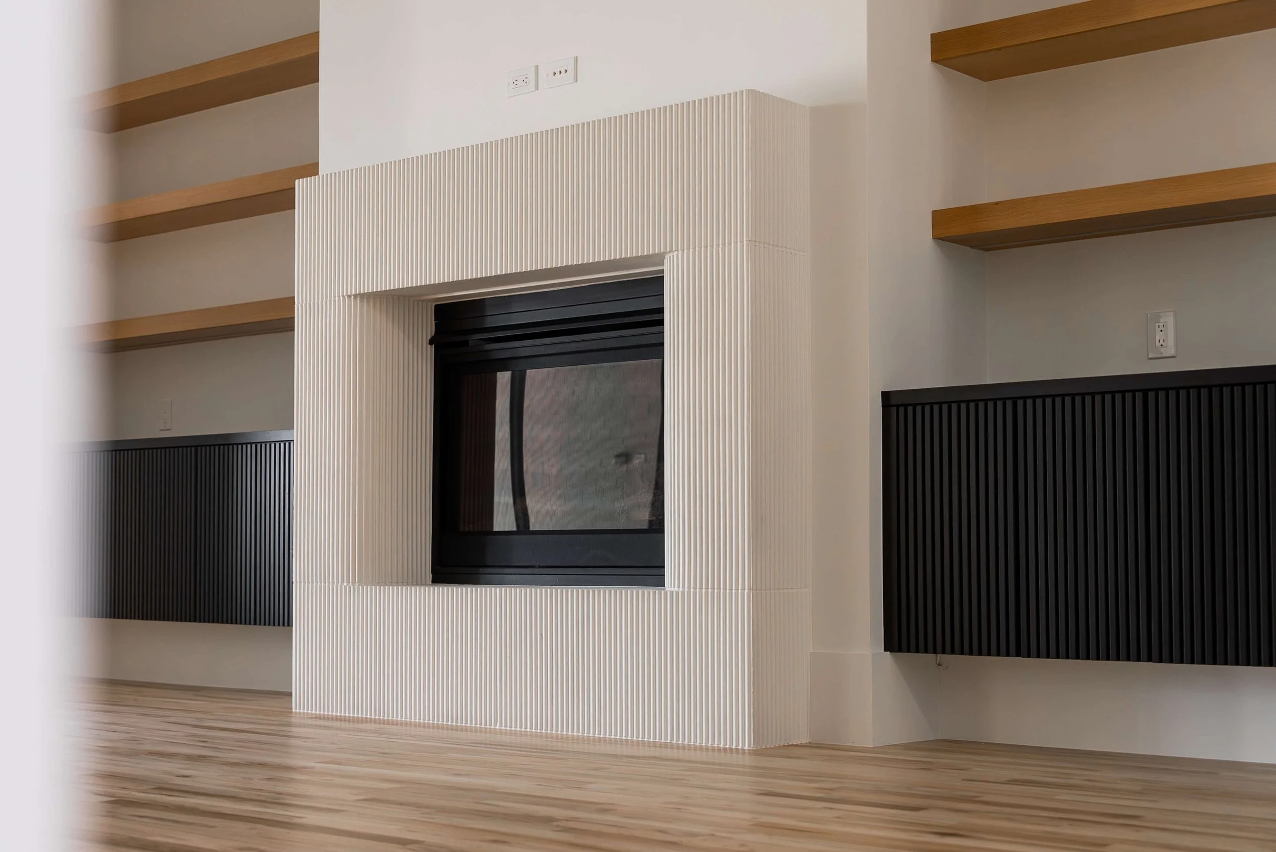 Modern living room fireplace with textured white surround and black glass door, flanked by black wall panels and wooden shelves on the wall.