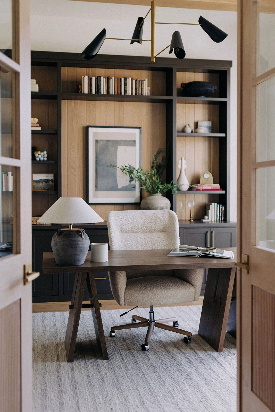 Home office with a wooden desk, beige office chair, bookshelf with books and decorative items, table lamp, and framed artwork on the wall.