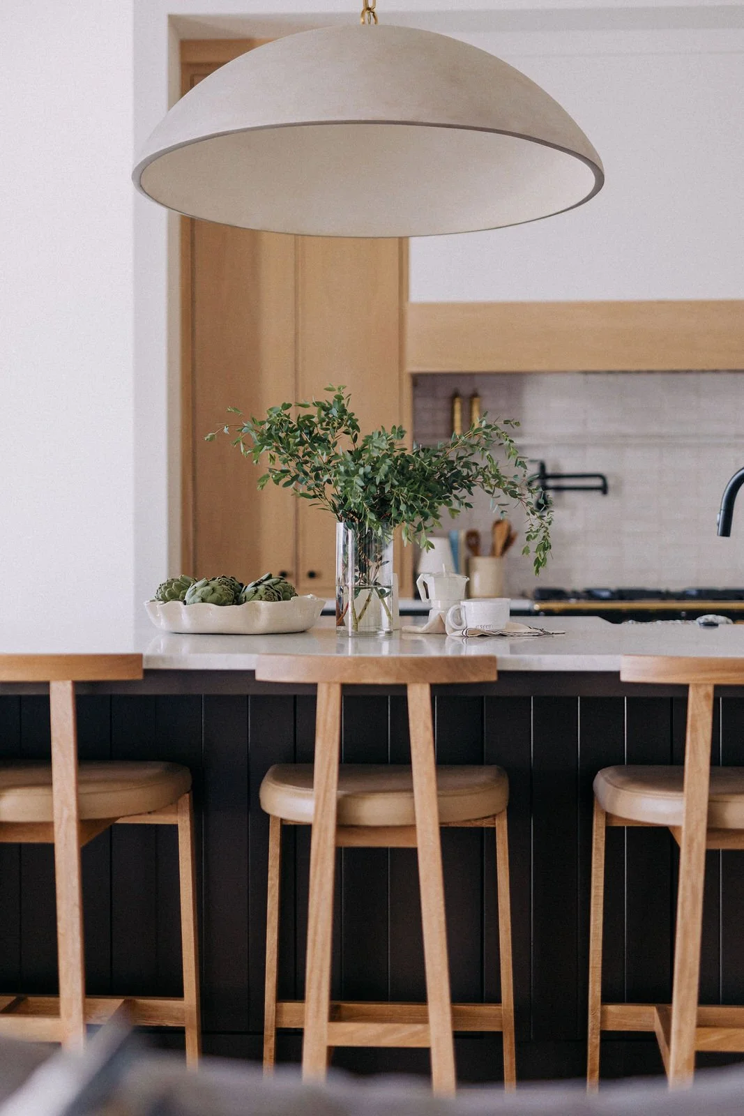 A kitchen island with a white countertop, black base, three wooden bar stools, a bowl of artichokes, a glass vase with greenery, and folded towels.