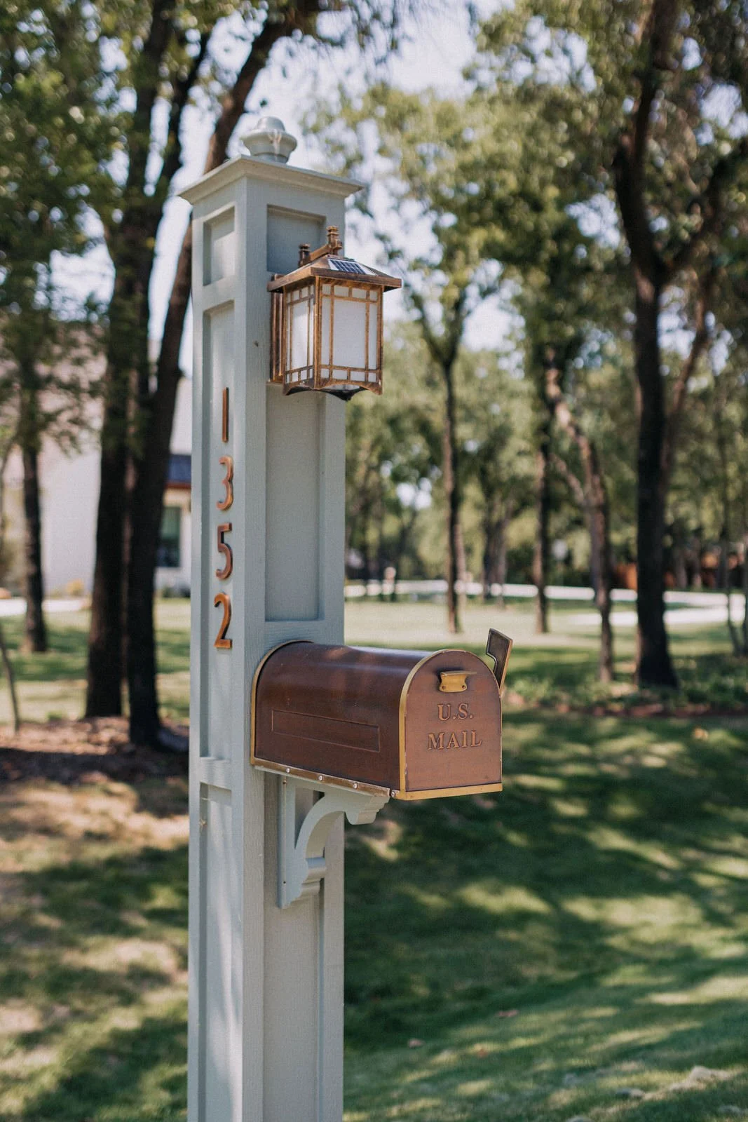 A white wooden mailbox post with the house number 1352 in gold letters, a vintage-style lantern, and a brown mailbox labeled 'U.S. MAIL' set in a green yard with trees.