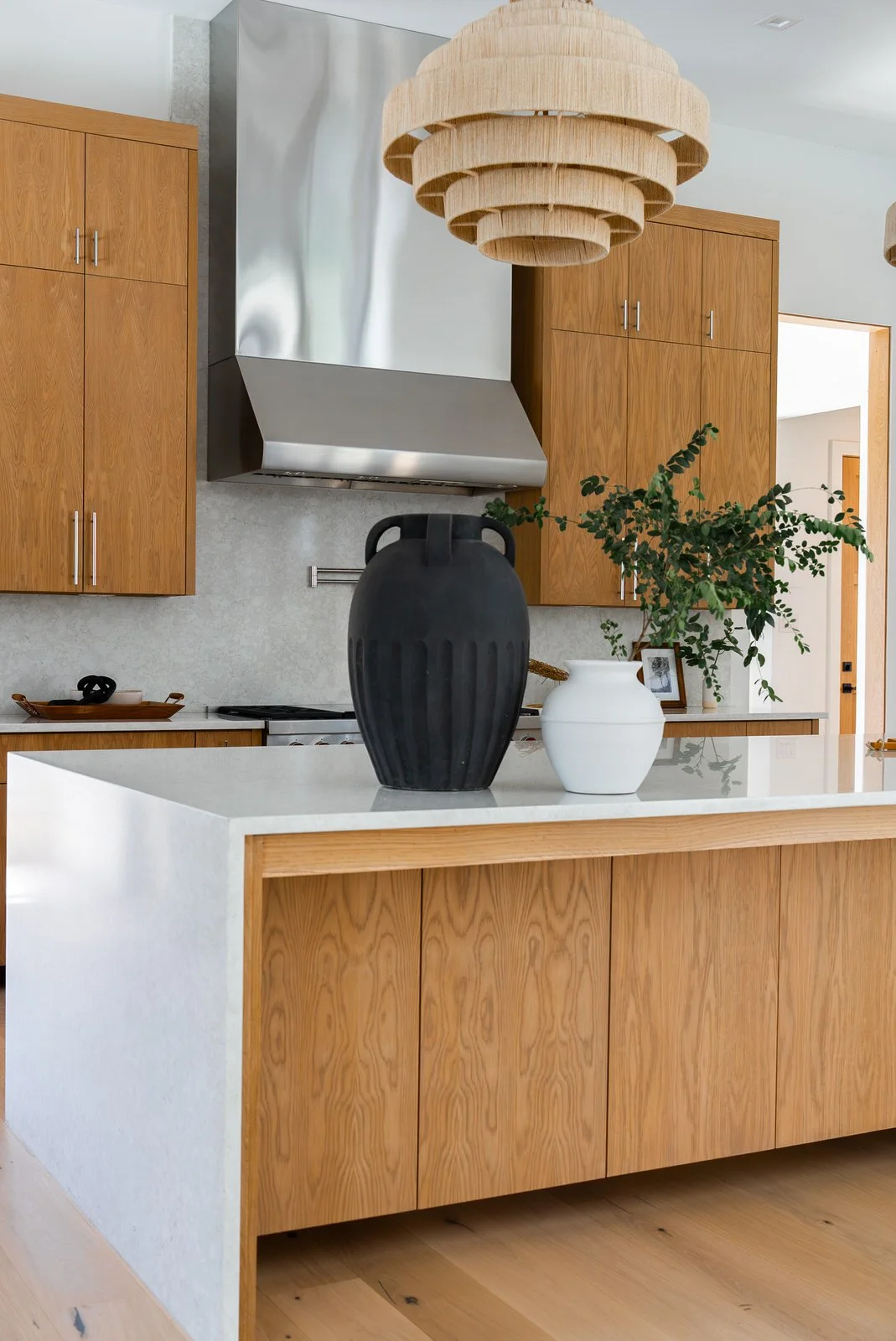 Modern kitchen with wooden cabinets, a white countertop island, black and white vases, a potted plant, a wooden pendant light, and a stainless steel range hood.