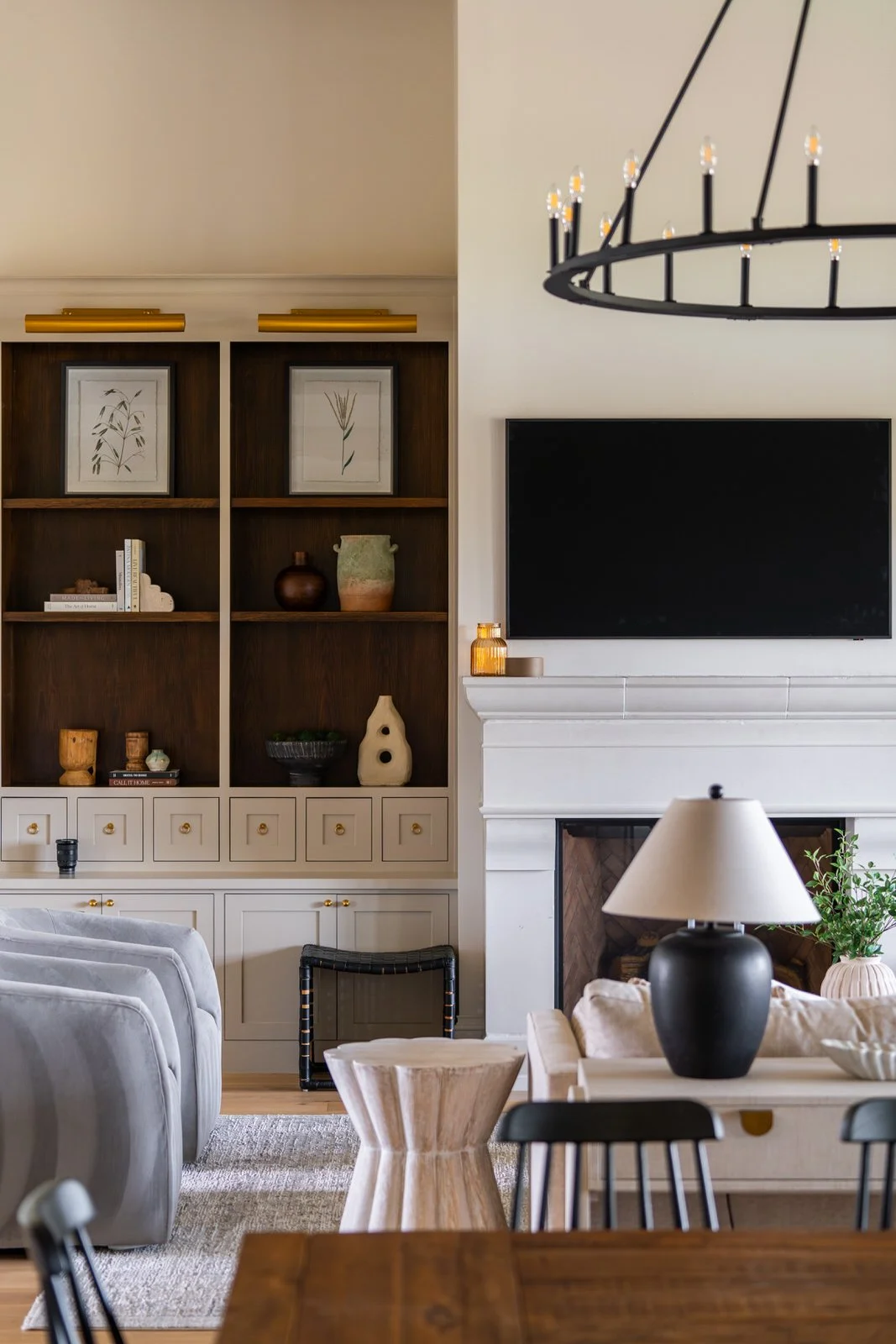 Living room with a built-in wooden bookshelf, a flat-screen TV above a white fireplace, a black chandelier, and a table with a lamp, plants, and chairs.