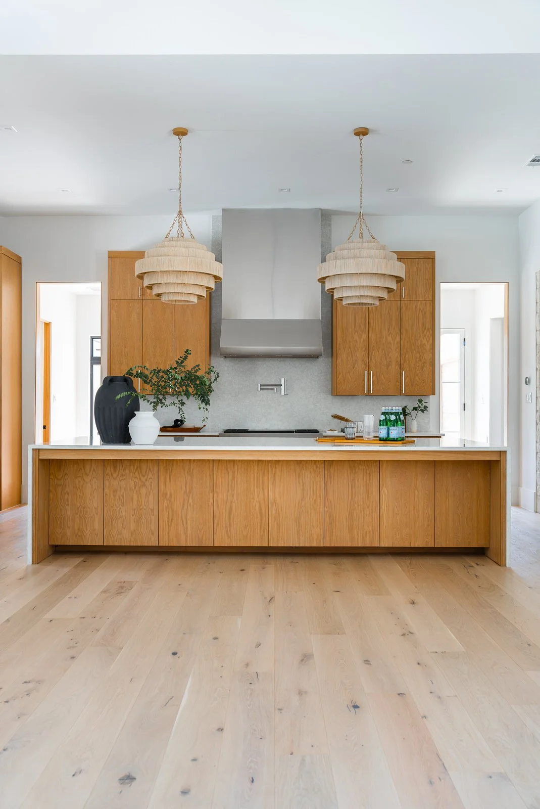 Modern kitchen with wooden cabinets, white countertop, two large pendant lights, stainless steel range hood, and light hardwood flooring.
