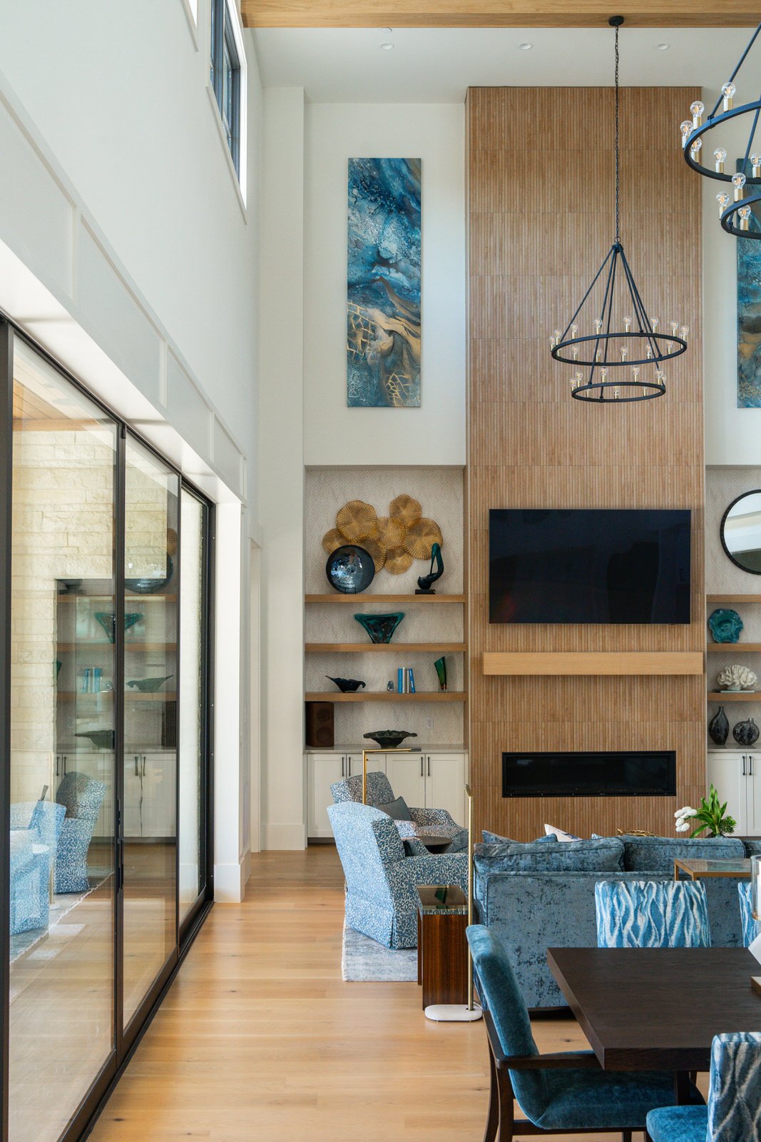 Interior view of a living room with a high ceiling, featuring a wood-paneled fireplace wall, a mounted flat-screen TV, built-in shelves with decorative objects, a modern chandelier, and large windows with sliding glass doors.