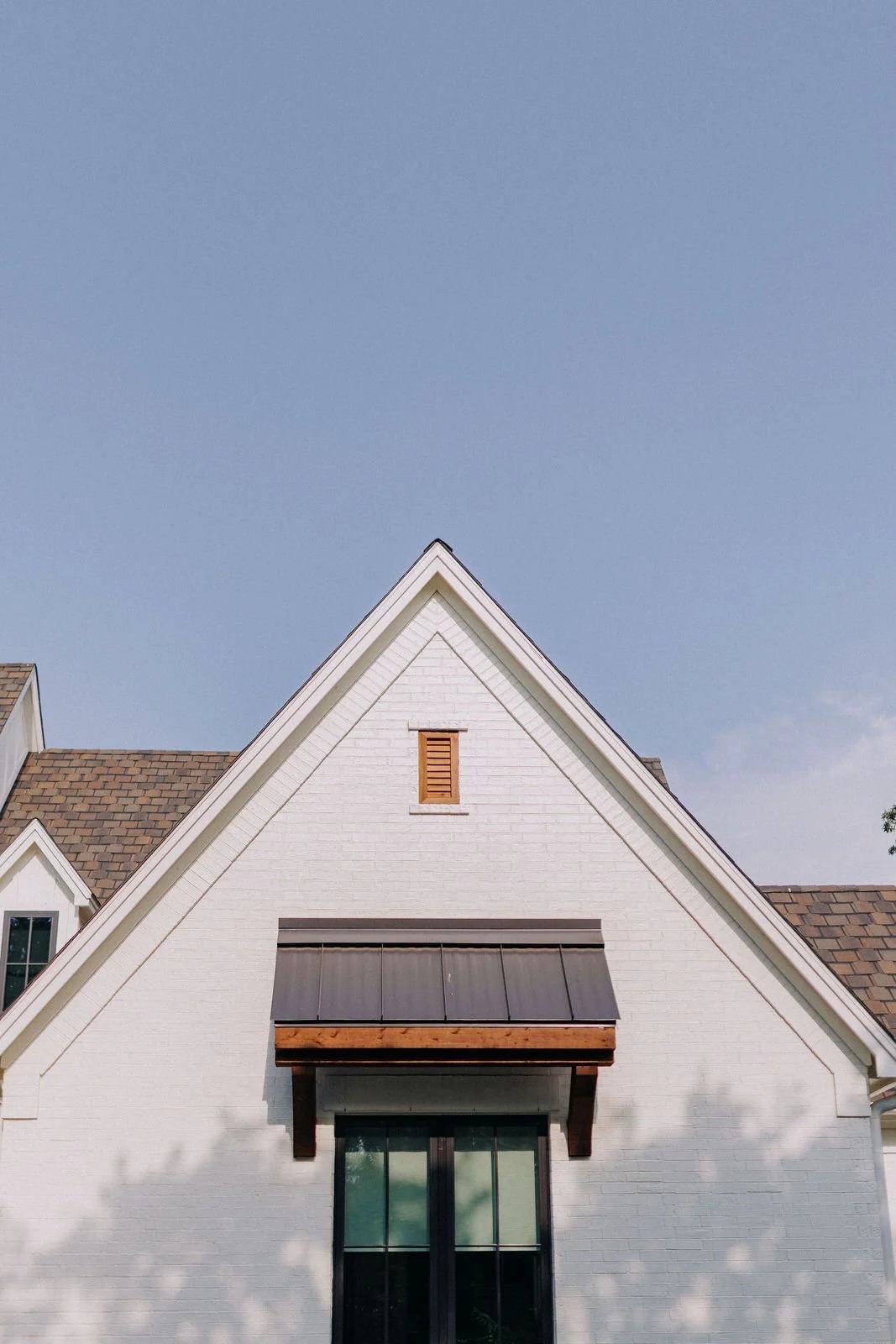 Luxury custom home - White house with brown shingle roof, small window with wooden shutter, and a large window with a wooden awning, under a clear blue sky.