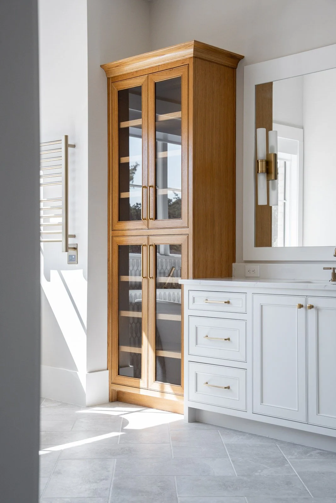 A bathroom corner featuring a white vanity with gold hardware, a large mirror, a light fixture, and a wooden display cabinet with glass doors, sunlight streaming through a window.