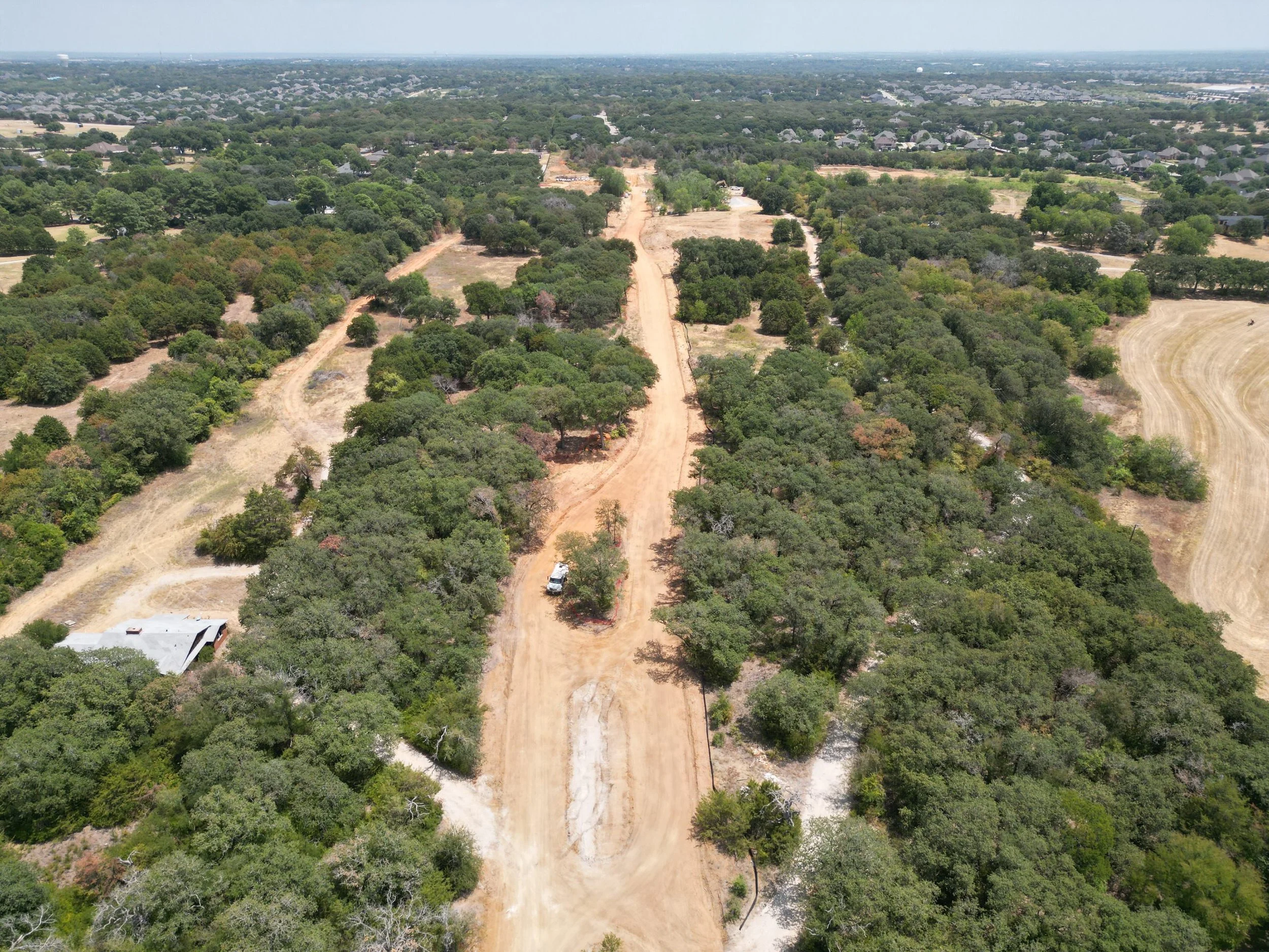 Aerial view of a dirt road construction site through a wooded area with trees and some open land, with residential neighborhoods visible in the distance.