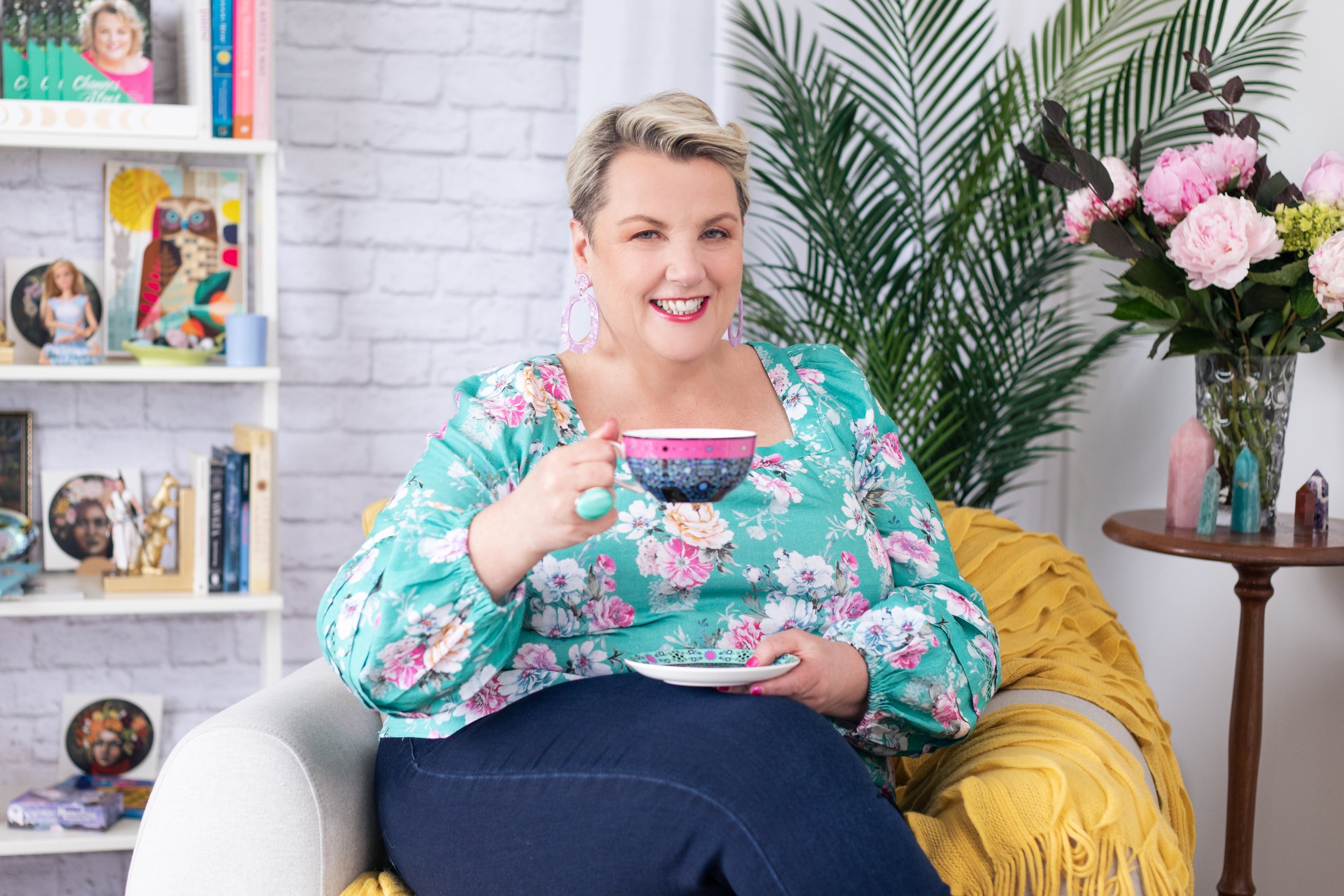 A smiling woman with short, blonde hair sitting on a light-colored chair, holding a colorful teacup with a matching saucer, in a cozy, well-decorated room with white brick walls, bookshelves, large green plants, and a vase of pink flowers.