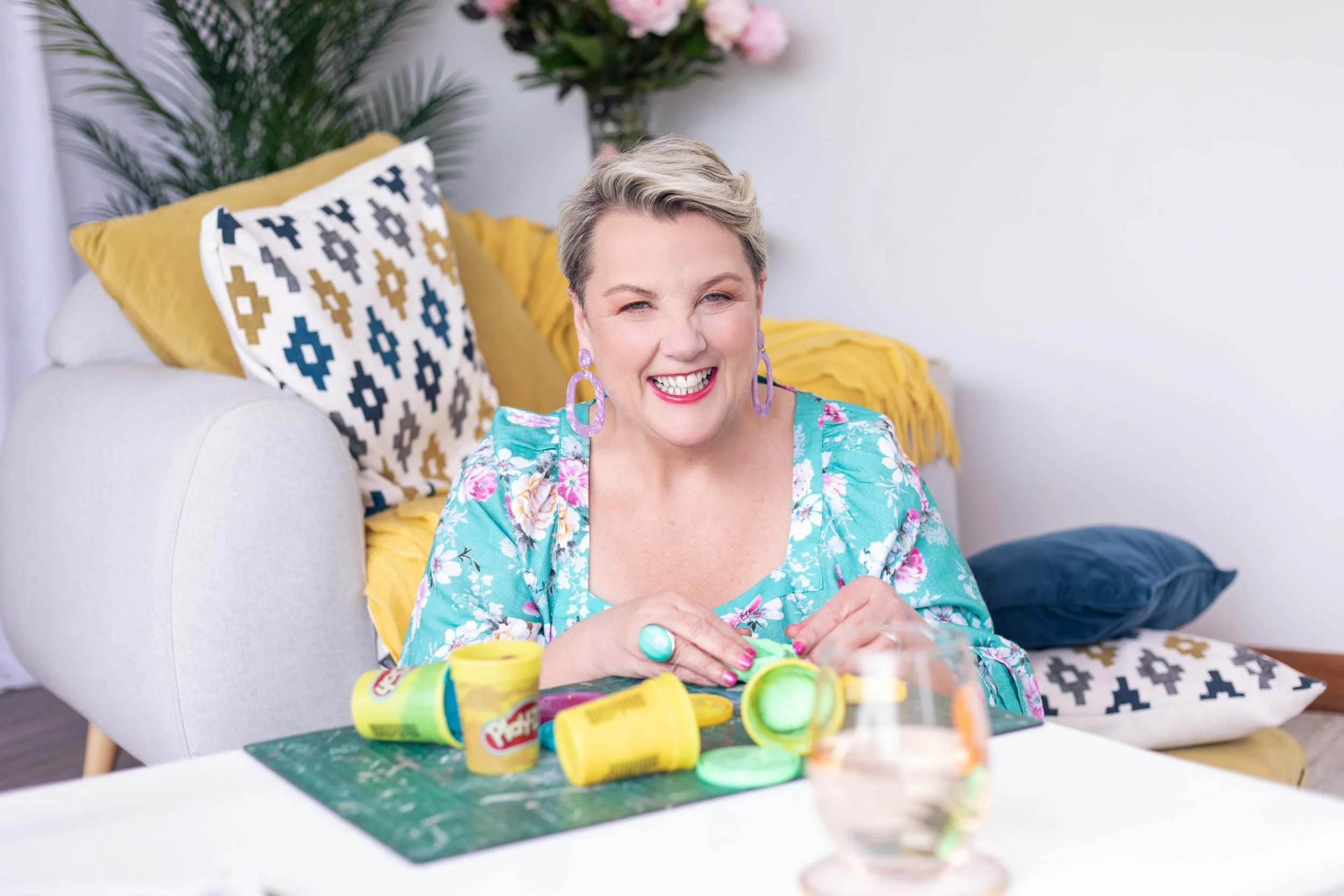 A smiling woman with short blonde hair, wearing purple earrings and a turquoise floral top, sitting at a table with Play-Doh containers in front of her, in a room with colorful pillows and a bouquet of pink flowers in the background.