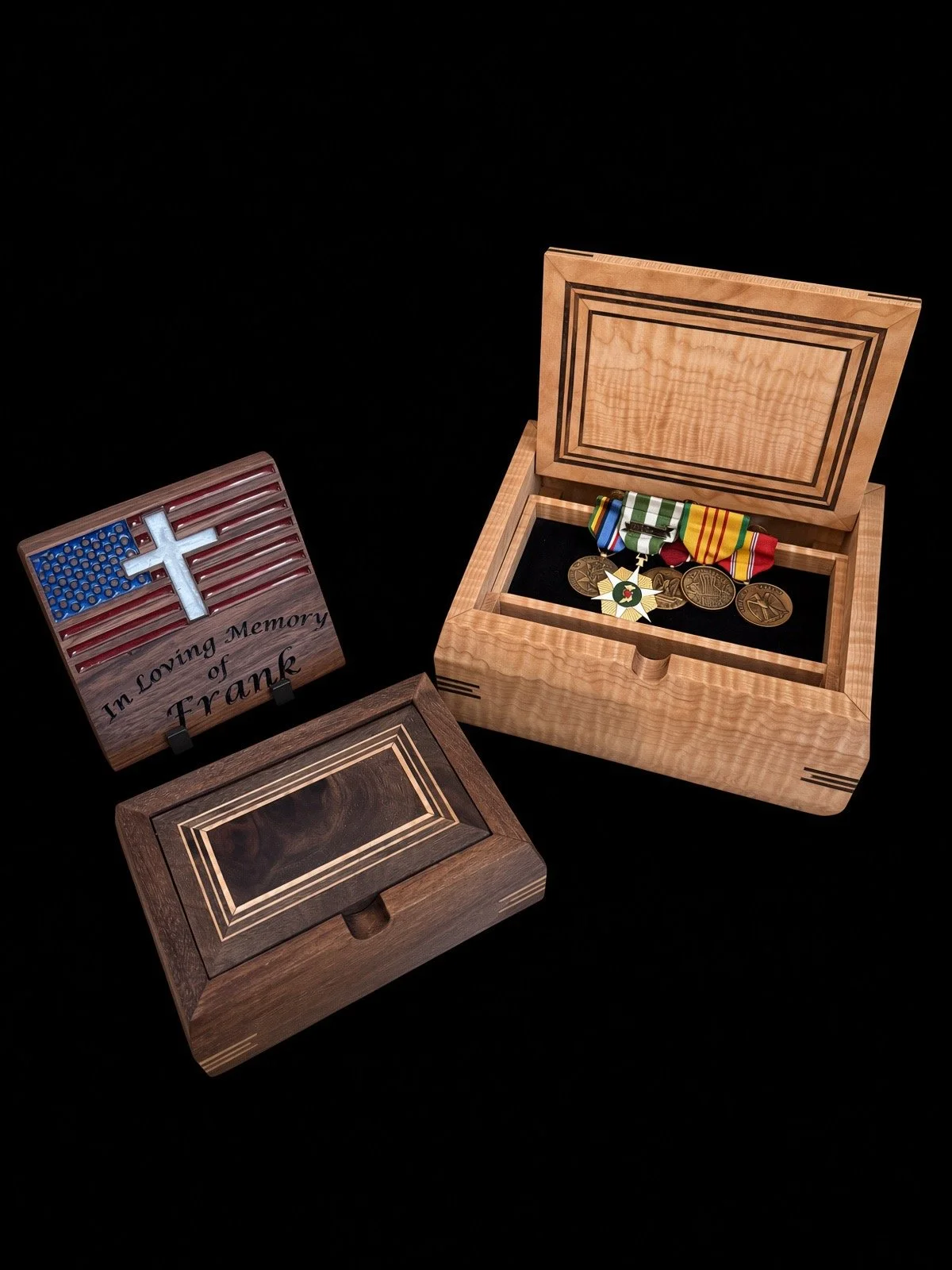 A wooden box with a lid containing military medals and ribbons, a small plaque with an American flag and cross and the inscription 'In Loving Memory of Frank,' and a separate wooden cover with decorative borders.
