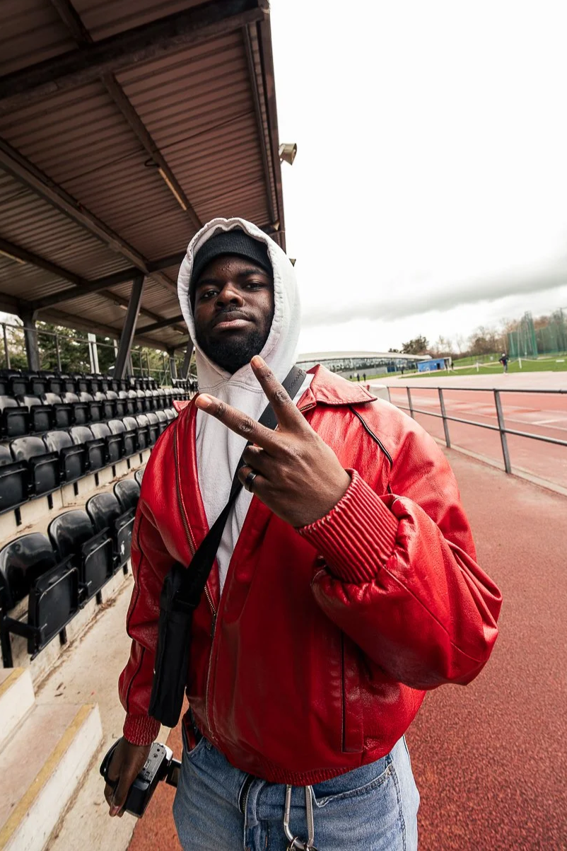 A man wearing a red jacket, gray hoodie, and black beanie making a peace sign with his fingers at an outdoor sports track and stadium, overcast sky in background.