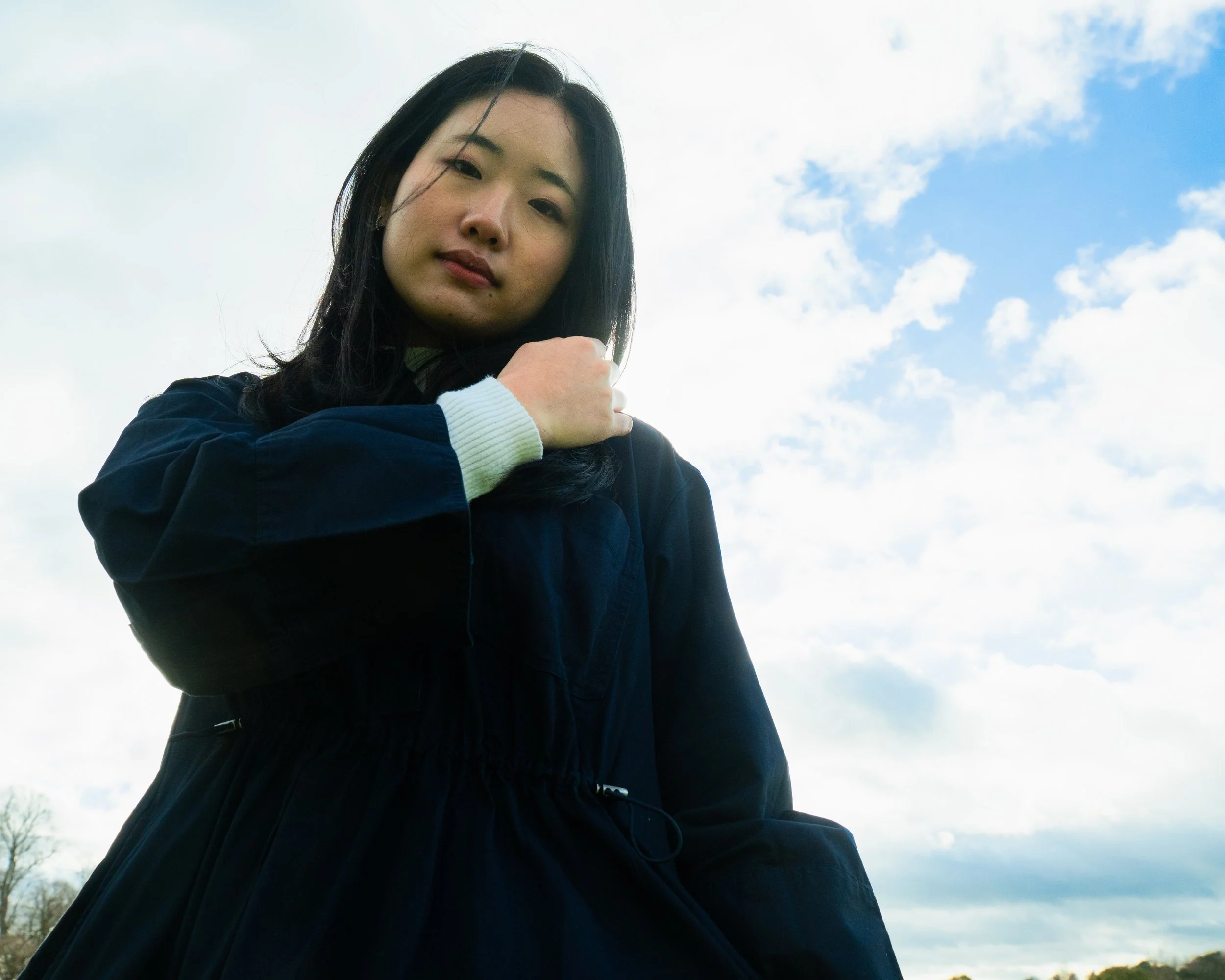 A young woman with long dark hair wearing a navy jacket and a white sweater, standing outdoors against a cloudy sky.