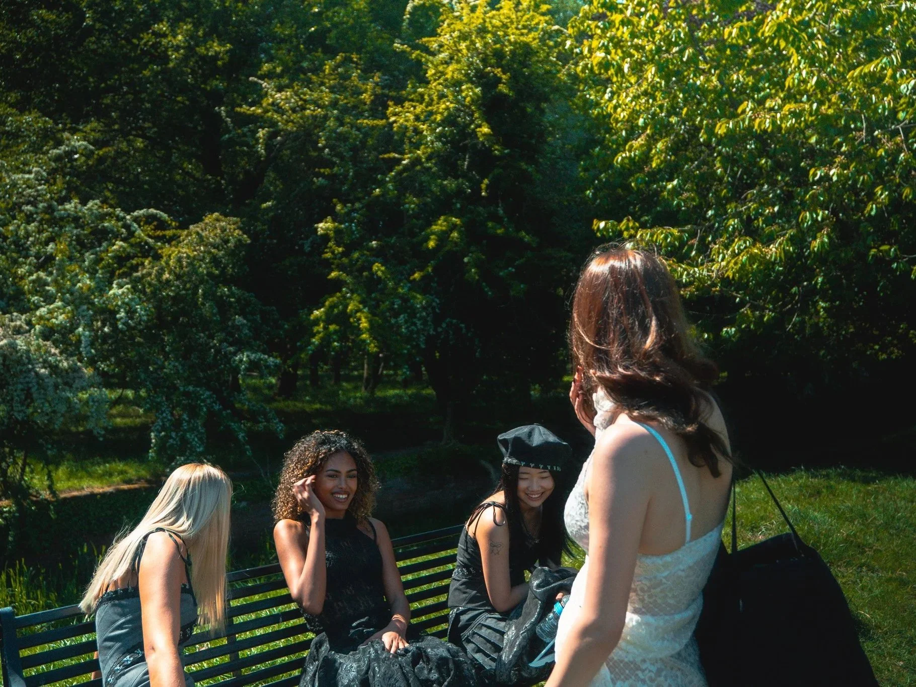 Four young women sitting and standing on a park bench surrounded by green trees, one woman in a graduation cap and gown.