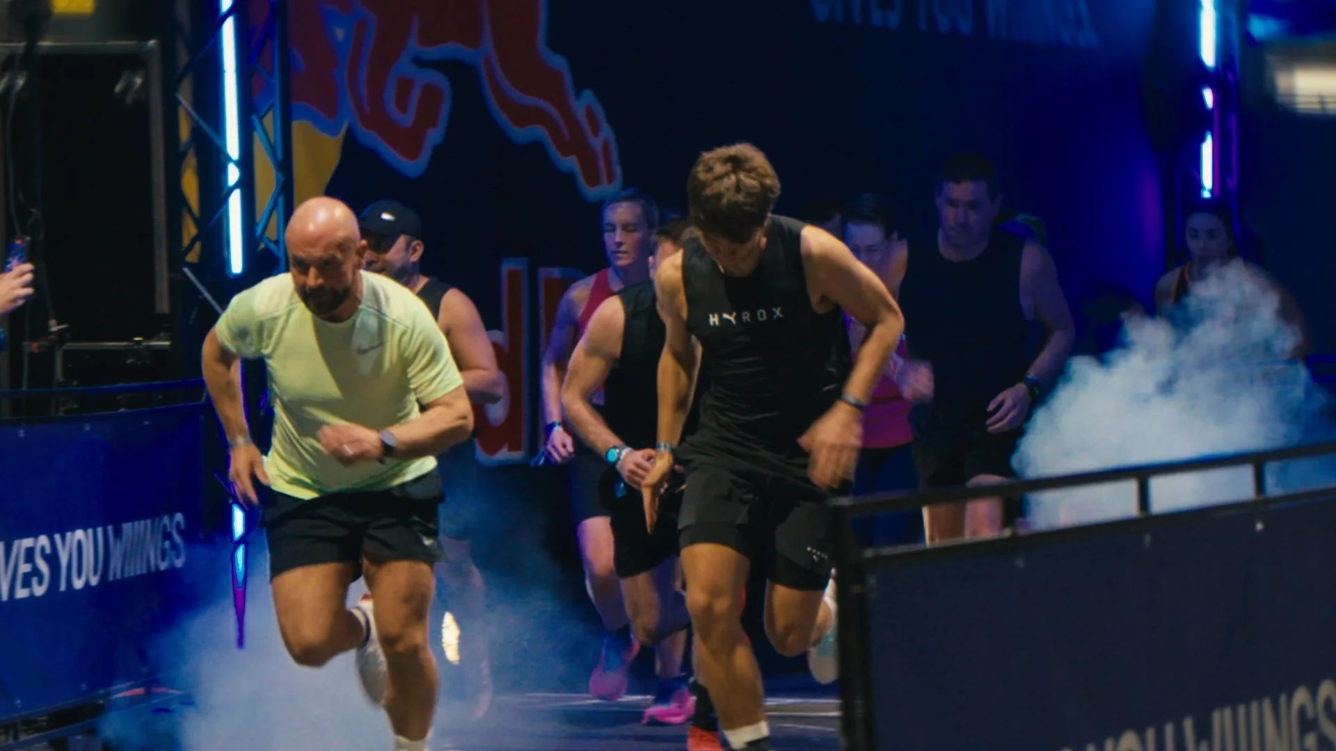 A group of people running in an indoor arena during a race or marathon event, with branding and lighting effects in the background.