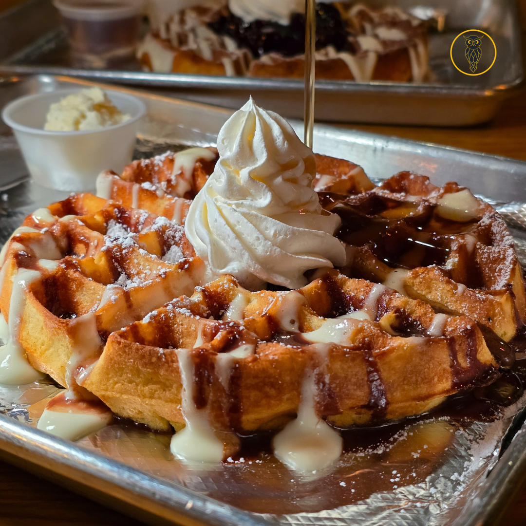 A waffle topped with whipped cream, drizzled with chocolate and caramel sauces, with extra whipped cream on the side. In the background, there is a tray with more waffles.