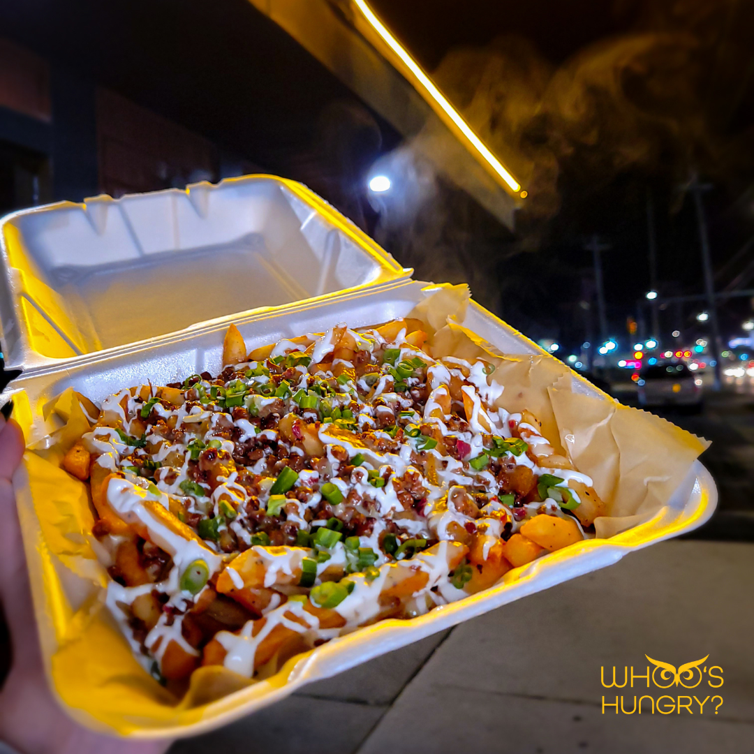 A takeout container filled with loaded fries topped with cheese, chopped chives, and drizzled with sauces, held outdoors at night with streetlights and cars visible in the background.