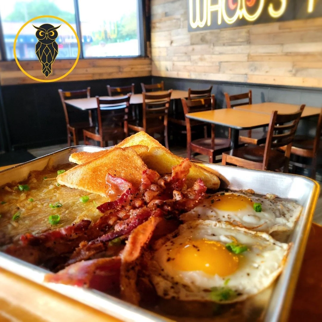 A breakfast dish with fried eggs, crispy bacon, toast, and hash browns served in a metal tray inside a restaurant with wooden walls and chairs.