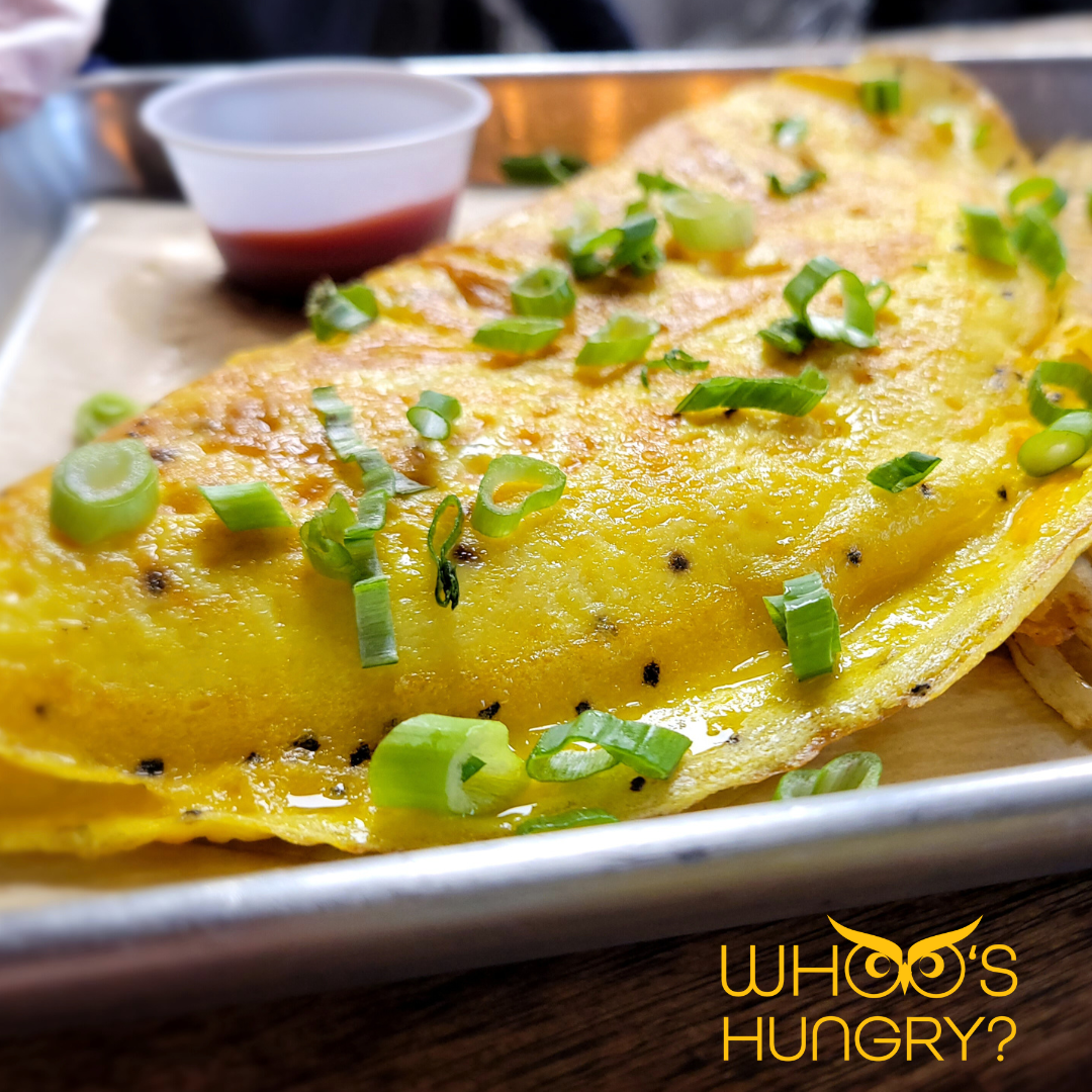 Close-up of a crispy, golden-yellow omelet garnished with chopped green onions, served on a metal tray with a side of red sauce in a small cup.