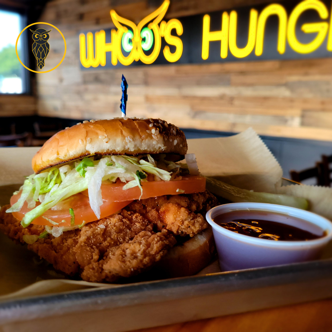 Fried chicken sandwich with lettuce, tomato, pickles, and a sesame seed bun, served with a small cup of dark soy-based dipping sauce on a tray in a restaurant with a wooden wall and neon sign reading 'WHO'S HUNG' in the background.
