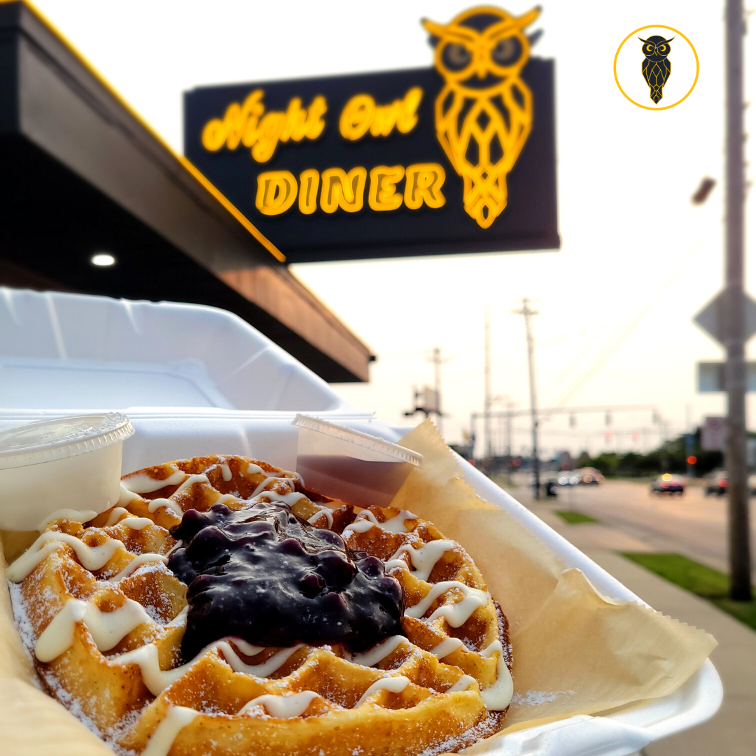 A waffle topped with blueberries and white icing, served with two small containers of syrup, in a takeout container outside a diner with illuminated sign reading "Night Owl Diner" and an owl logo.