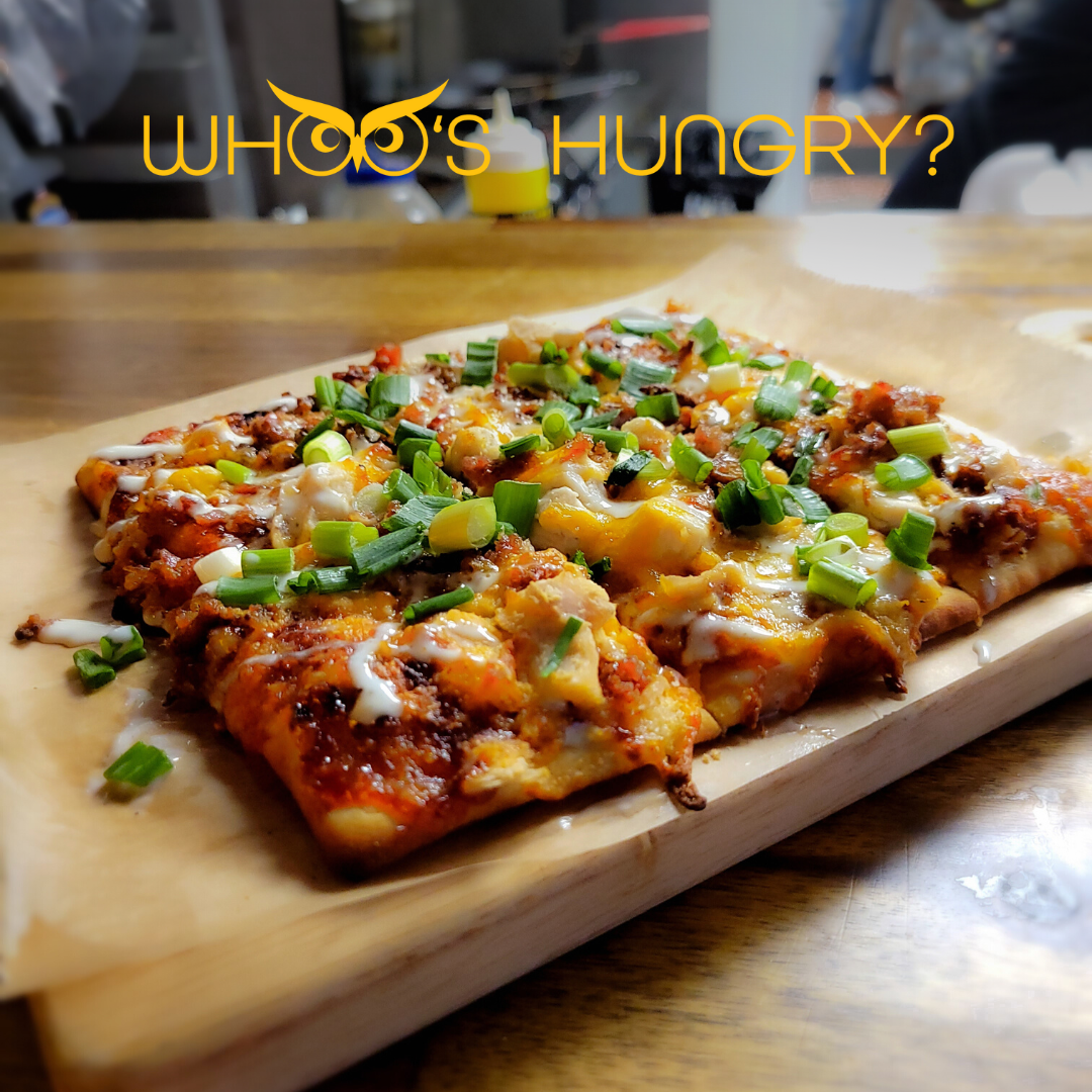 A close-up of a freshly baked pizza topped with green onions, melted cheese, and various toppings on a wooden serving board at a restaurant.