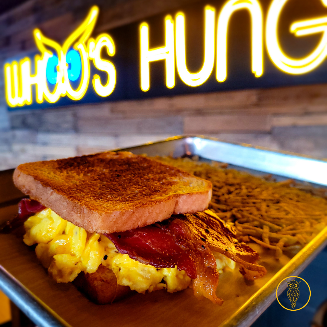 A breakfast sandwich with scrambled eggs, bacon, and a toasted bread slice, served on a tray with a side of crispy hash browns, in front of a bright yellow neon sign reading 'WHO'S HUNG'.