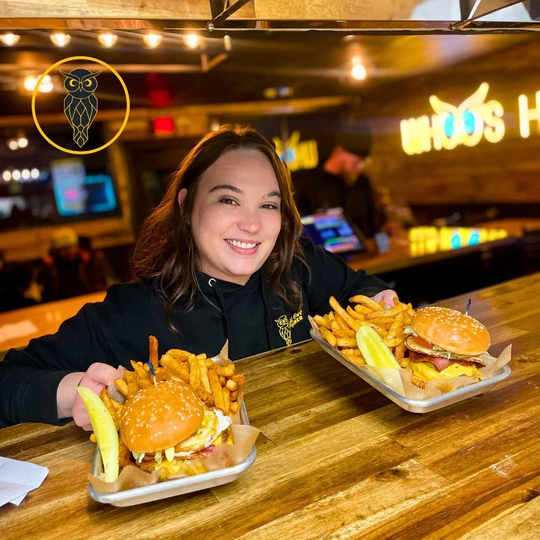A young woman smiling at the camera, sitting at a wooden bar counter with two trays of food in front of her. Each tray contains a burger topped with lettuce, tomato, cheese, and bacon, along with a side of French fries and a pickle spear. The backgro