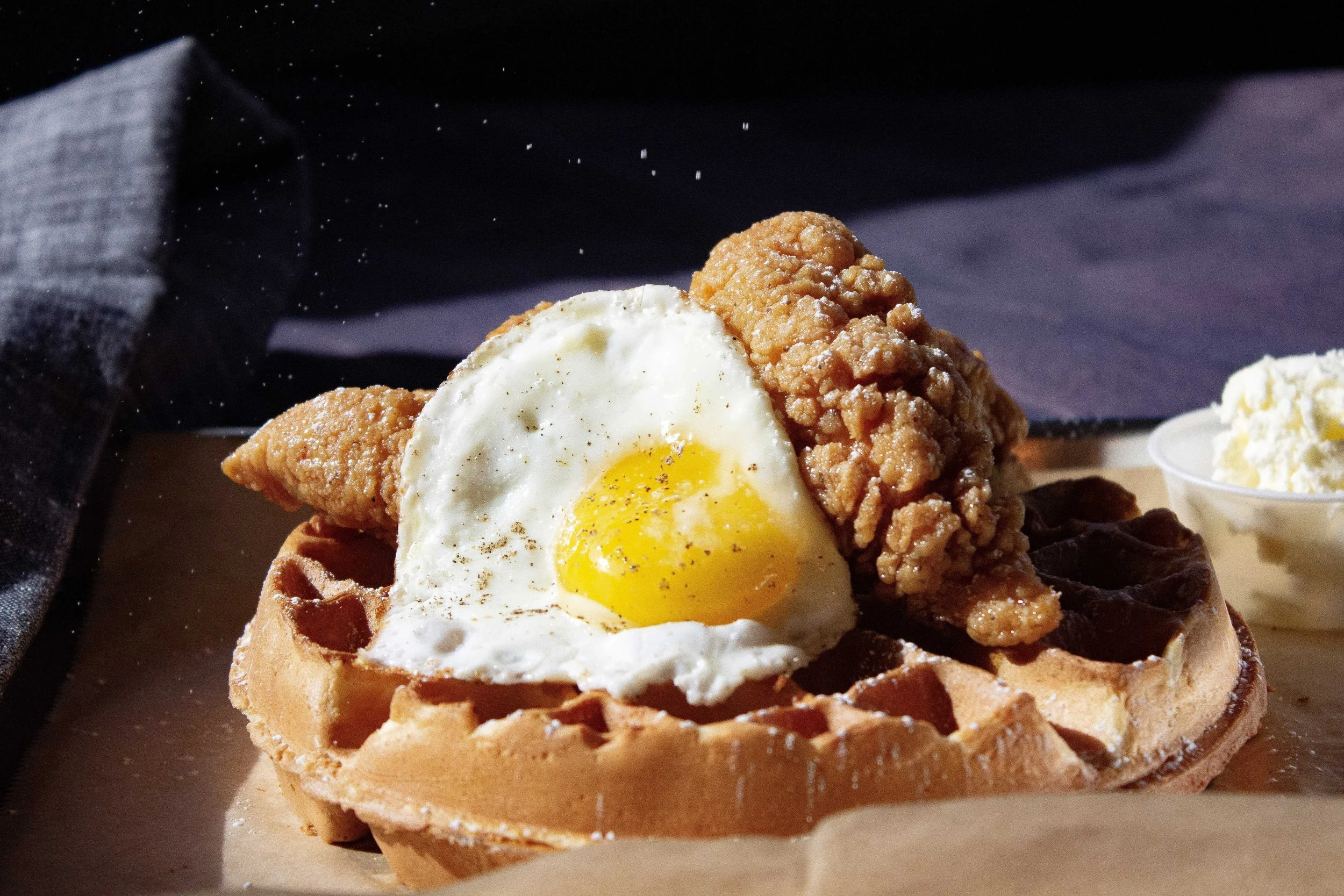 A waffle topped with a fried egg, fried chicken piece, and cinnamon sugar, served with a side of butter.
