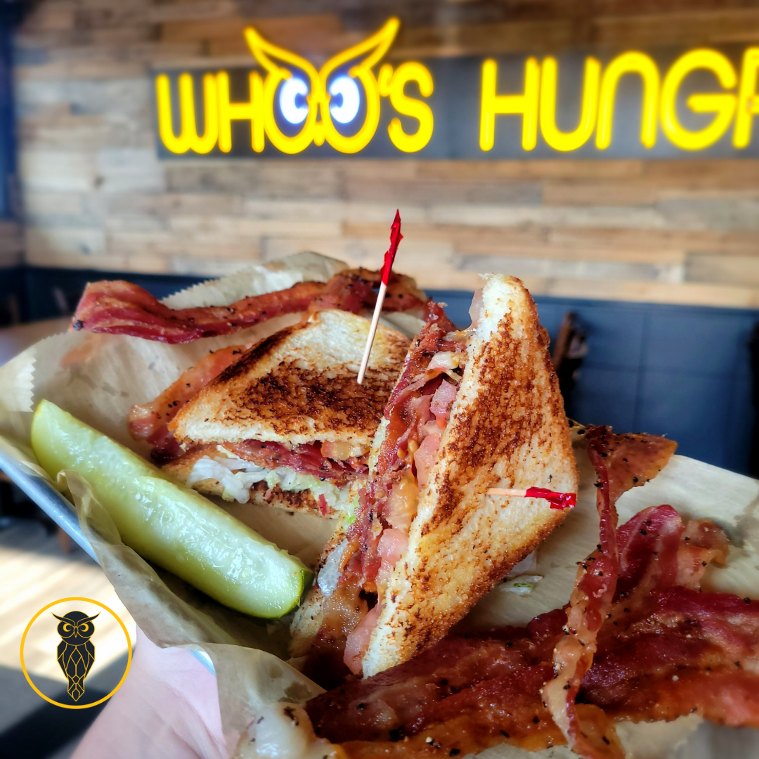A close-up of a BLT sandwich with crispy bacon, lettuce, tomato, and toasted bread, served with pickles, in a casual dining restaurant with a neon sign that reads 'WHO'S HUNGRY' in the background.