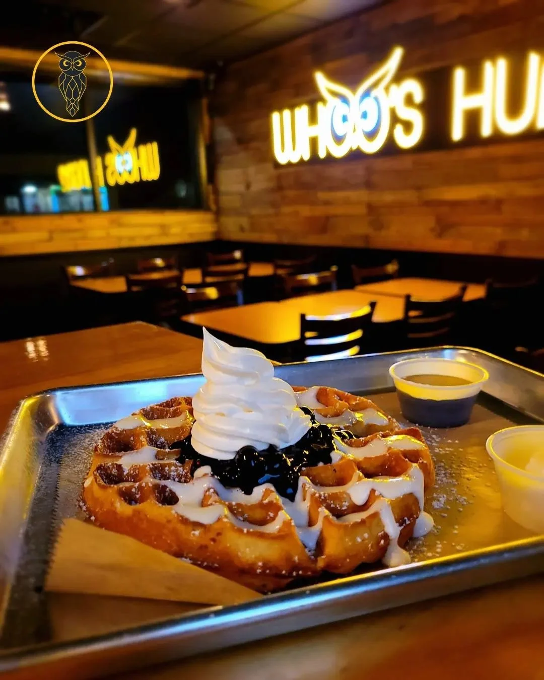 A waffle topped with whipped cream, chocolate syrup, and powdered sugar on a tray, with small containers of syrup on the side in a restaurant with wood-paneled walls and neon signs.
