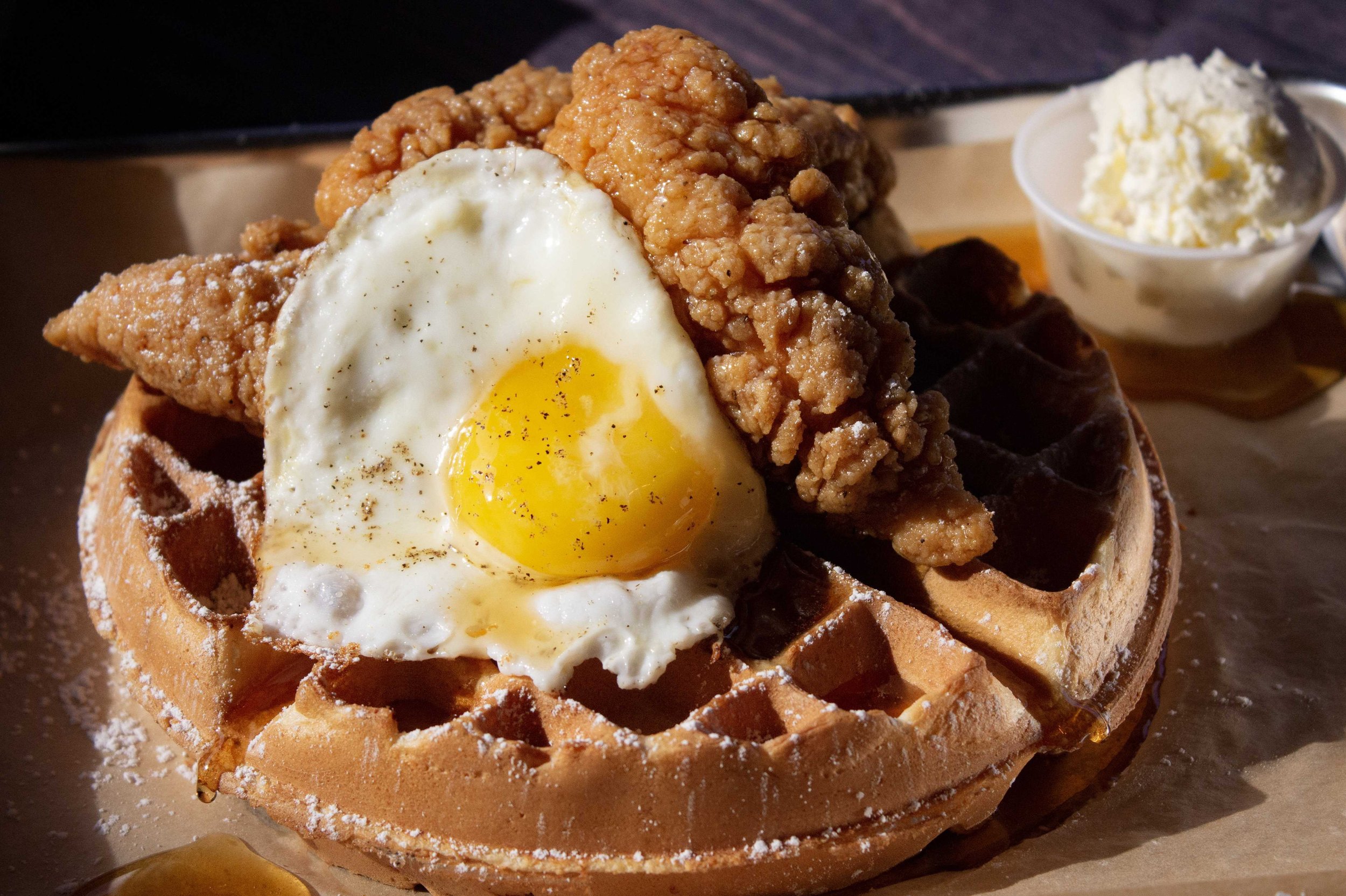 Waffle topped with fried chicken, sunny-side-up egg, and powdered sugar, served with a side of whipped butter.