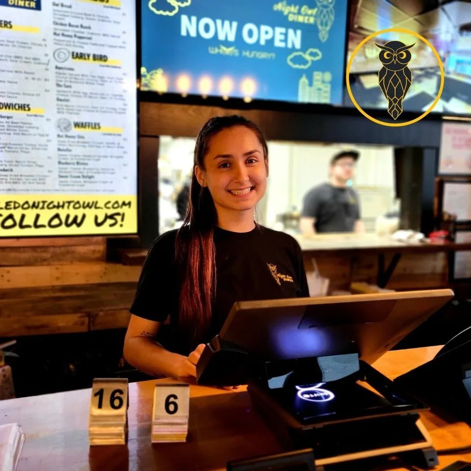 A young woman smiling at a counter in a restaurant, with a menu on the wall behind her, and a person blurred in the background. There is an owl logo in a yellow circle in the upper right corner.