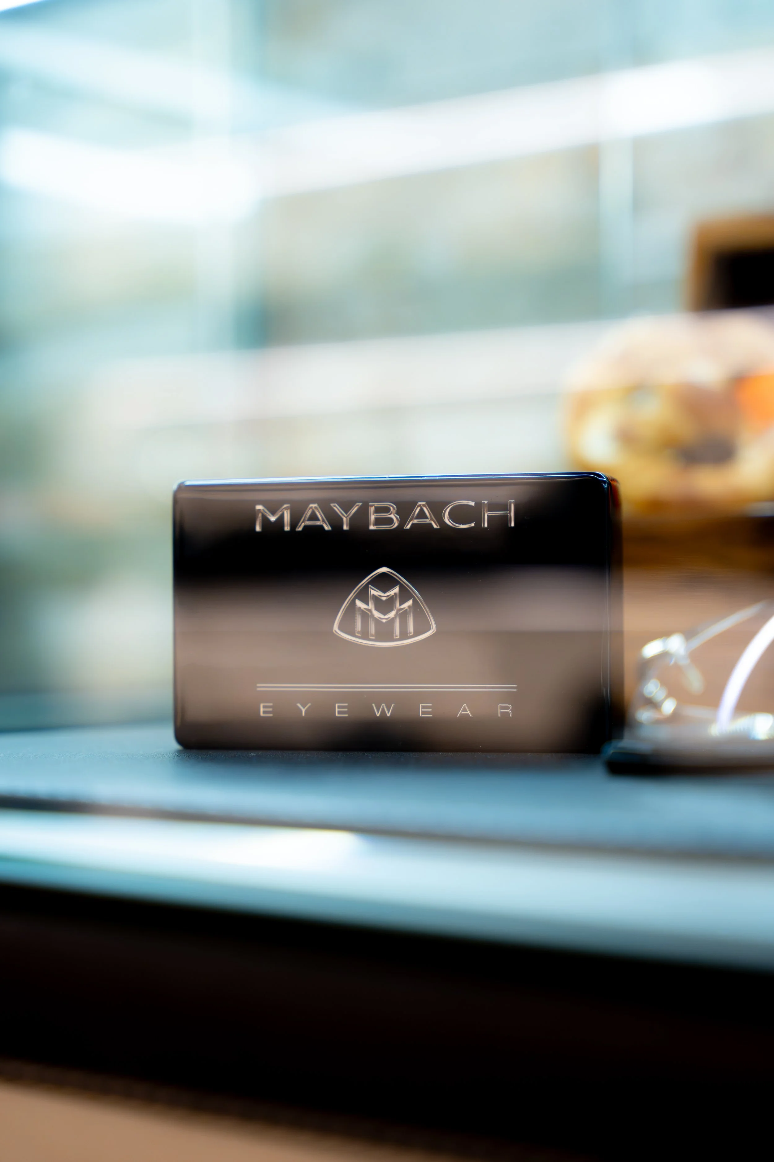 Nahaufnahme eines Maybach Eyewear-Produktes mit abgerundeter, schwarzer Verpackung und silberner Beschriftung, im Hintergrund unscharfe Szene mit Glasfenstern und Dekorationsgegenständen.